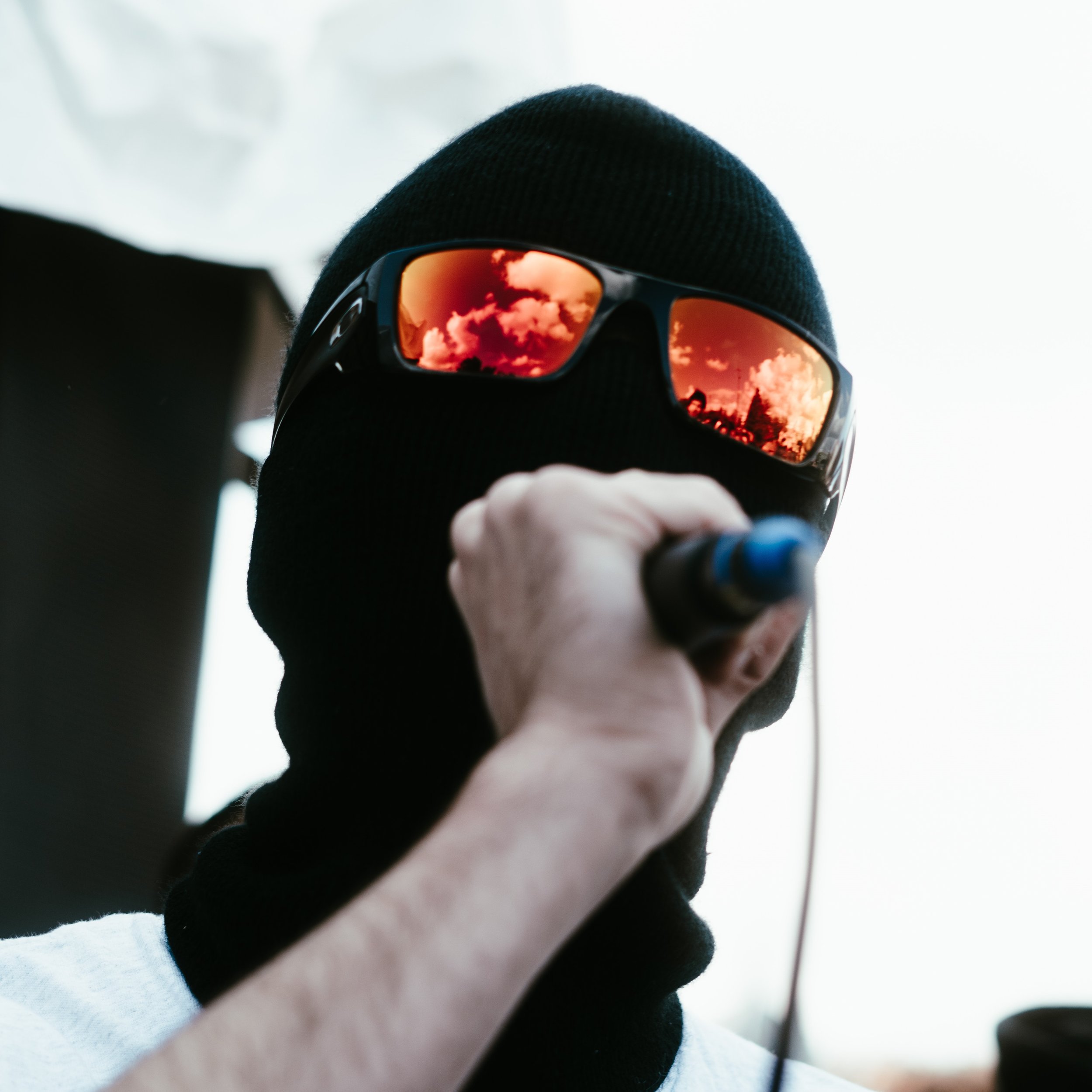 Personne portant un masque en tissu noir et des lunettes de soleil rouge-orange, tenant un objet noir, avec un fond flou.