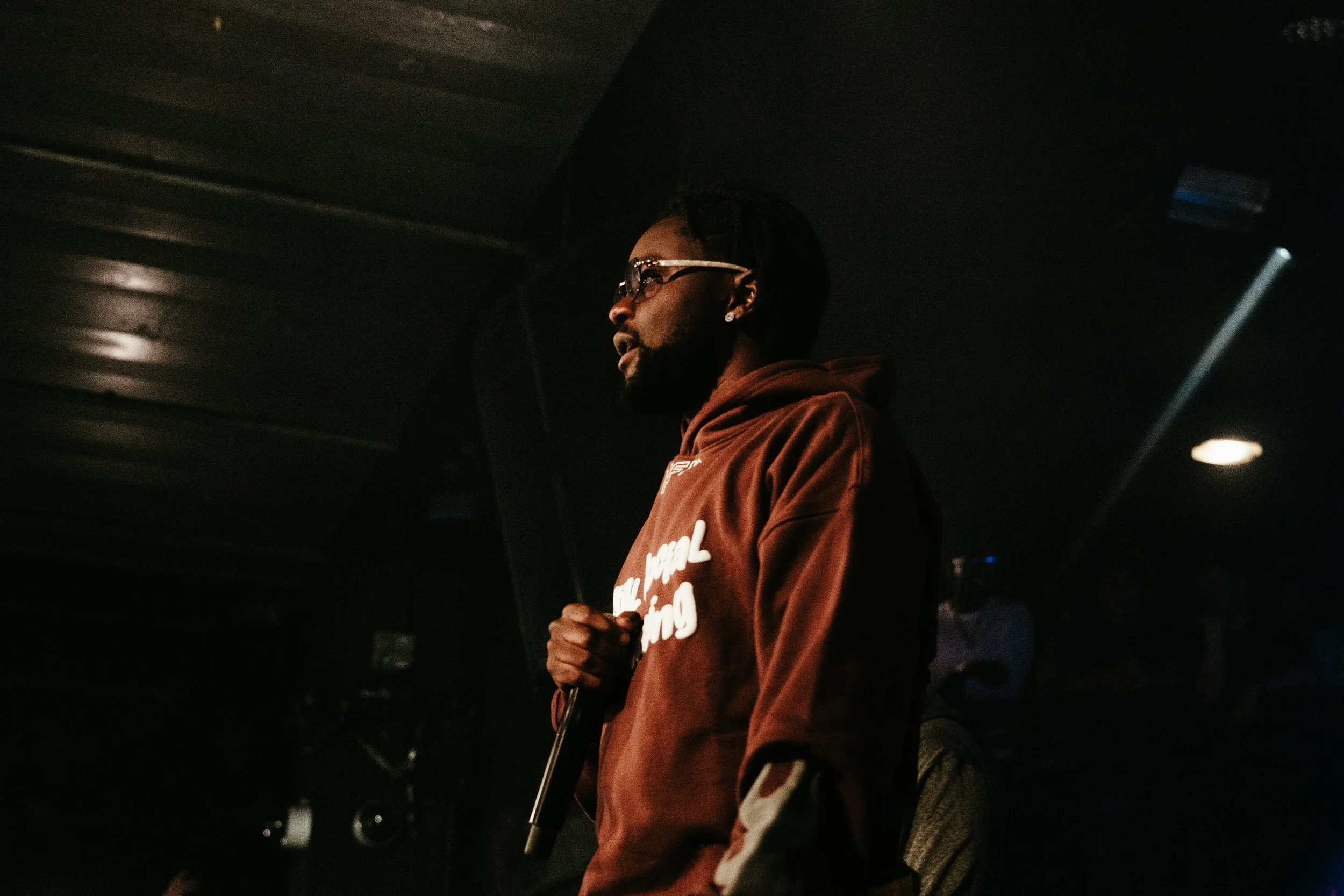 Un homme portant des lunettes, une barbe et une veste marron, tenant un microphone, dans une ambiance sombre et lumineuse