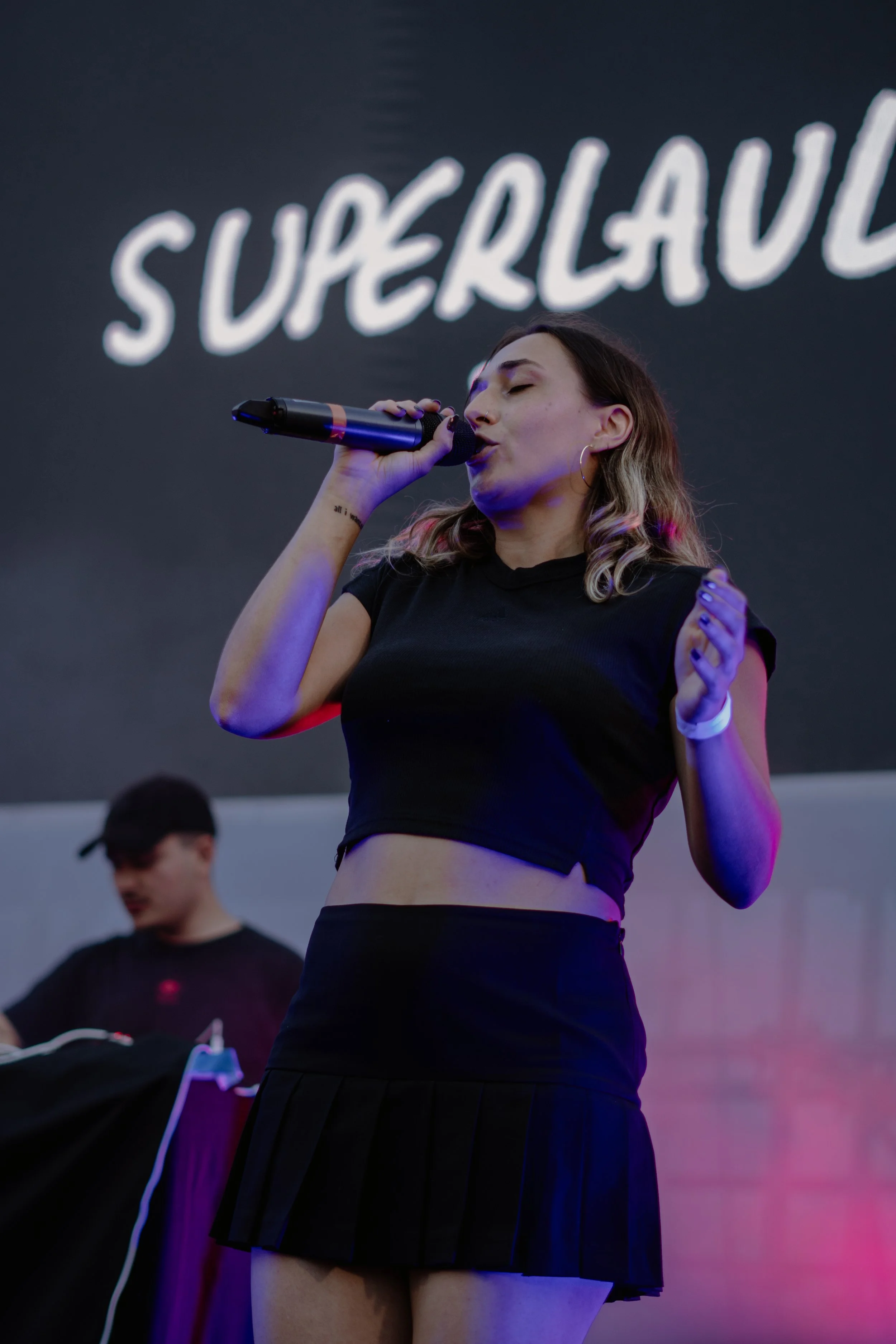 Une jeune femme chante dans un micro lors d'une performance, avec le mot 'SUPERLAVE' en arrière-plan.