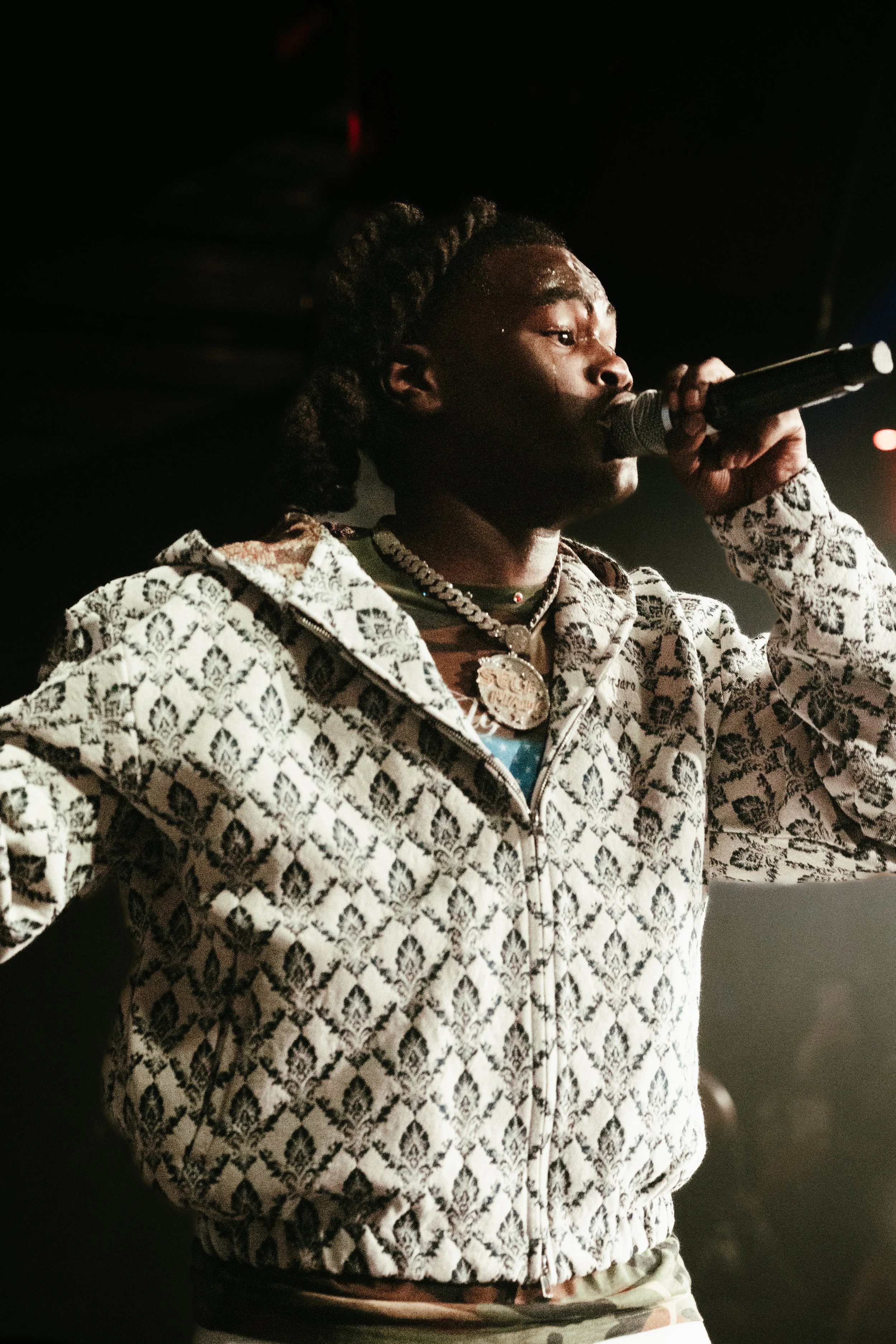 Un homme avec des dreadlocks tenant un microphone, portant un sweat à motifs, des colliers et des bijoux, en train de performer sur scène.