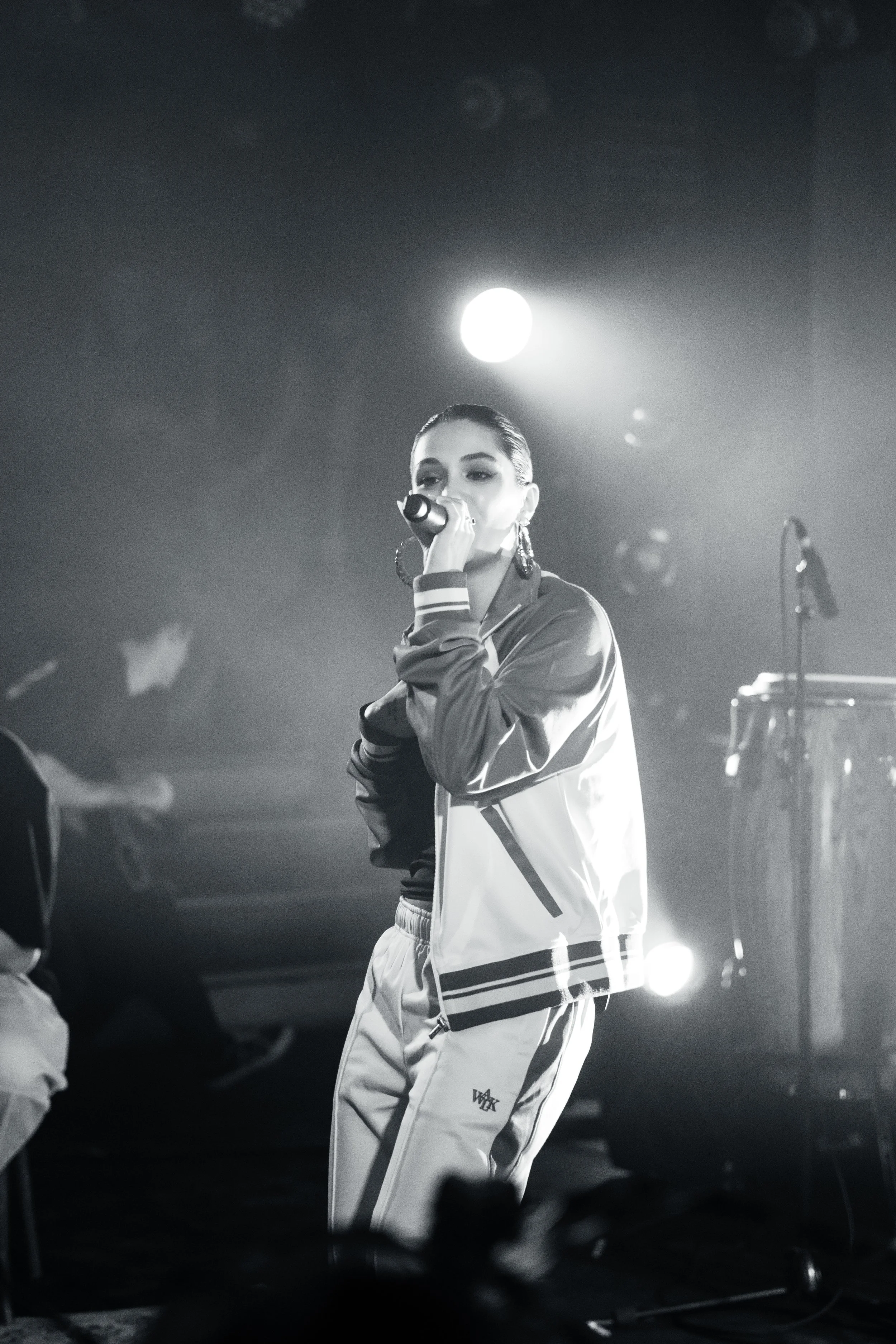 Une femme chante dans un micro lors d'une performance musicale sur scène, avec des lumières derrière elle.
