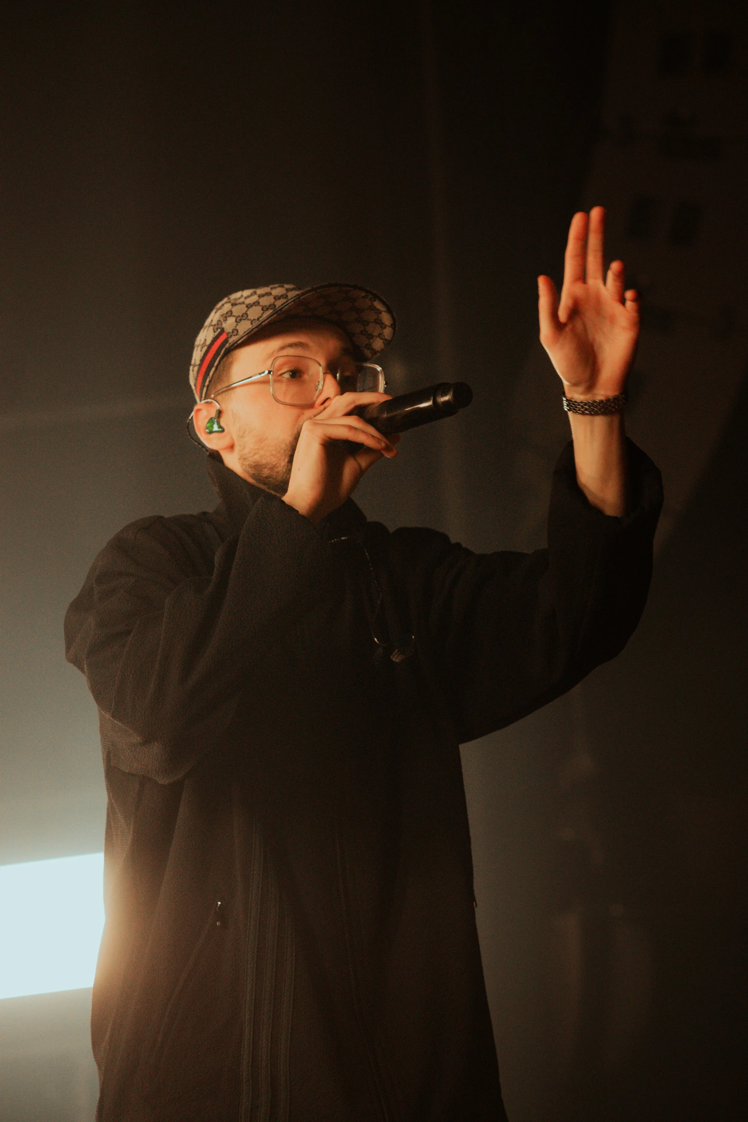 Un jeune homme avec des lunettes, un baseball cap Gucci, un écouteur vert, tenant un micro, en train de chanter ou de parler, avec un bras levé et la main ouverte, sur scène