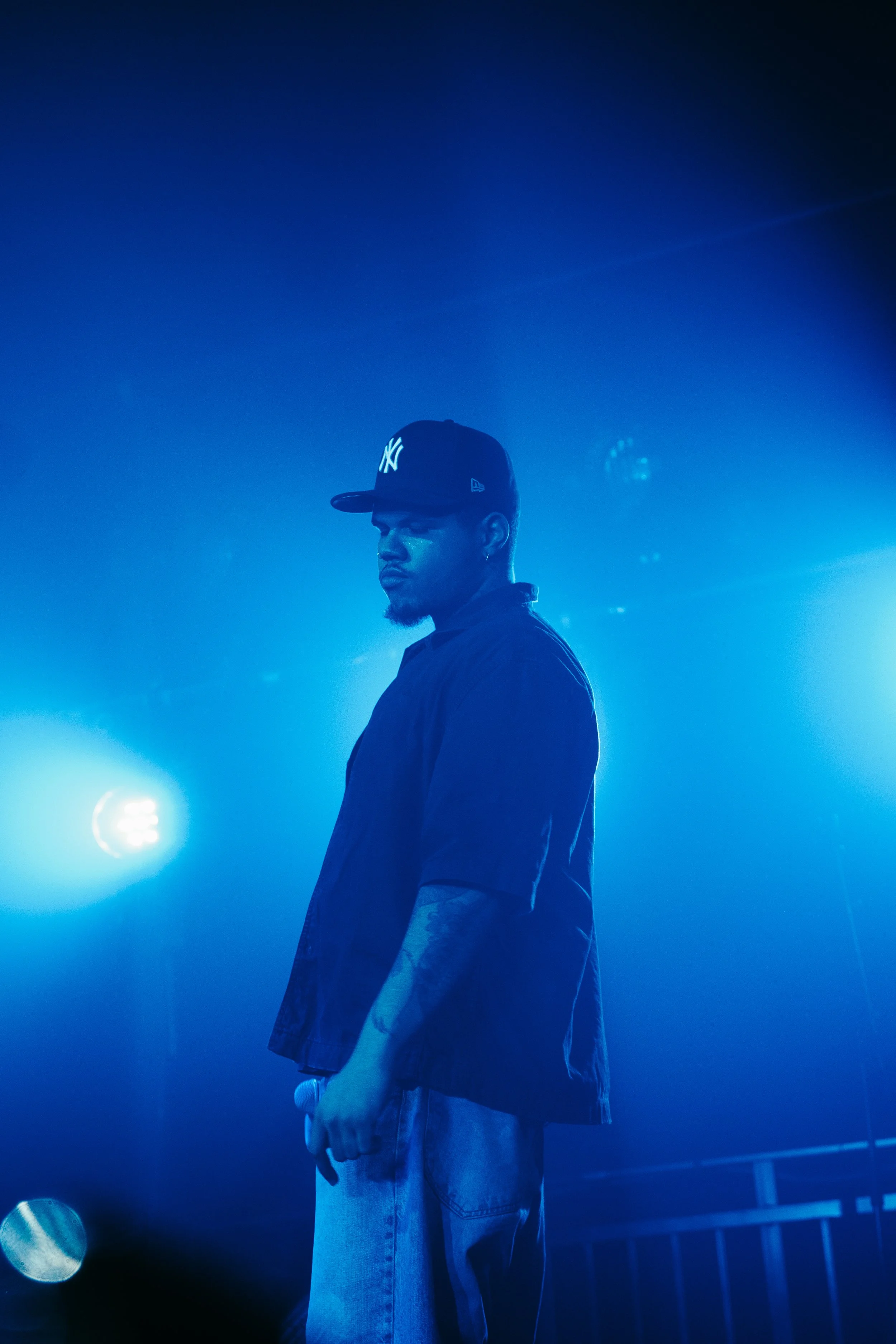 Un homme portant une casquette New York Yankees, debout sur scène avec un éclairage bleu, tenant un microphone dans la main, dans une ambiance sombre et mystérieuse.