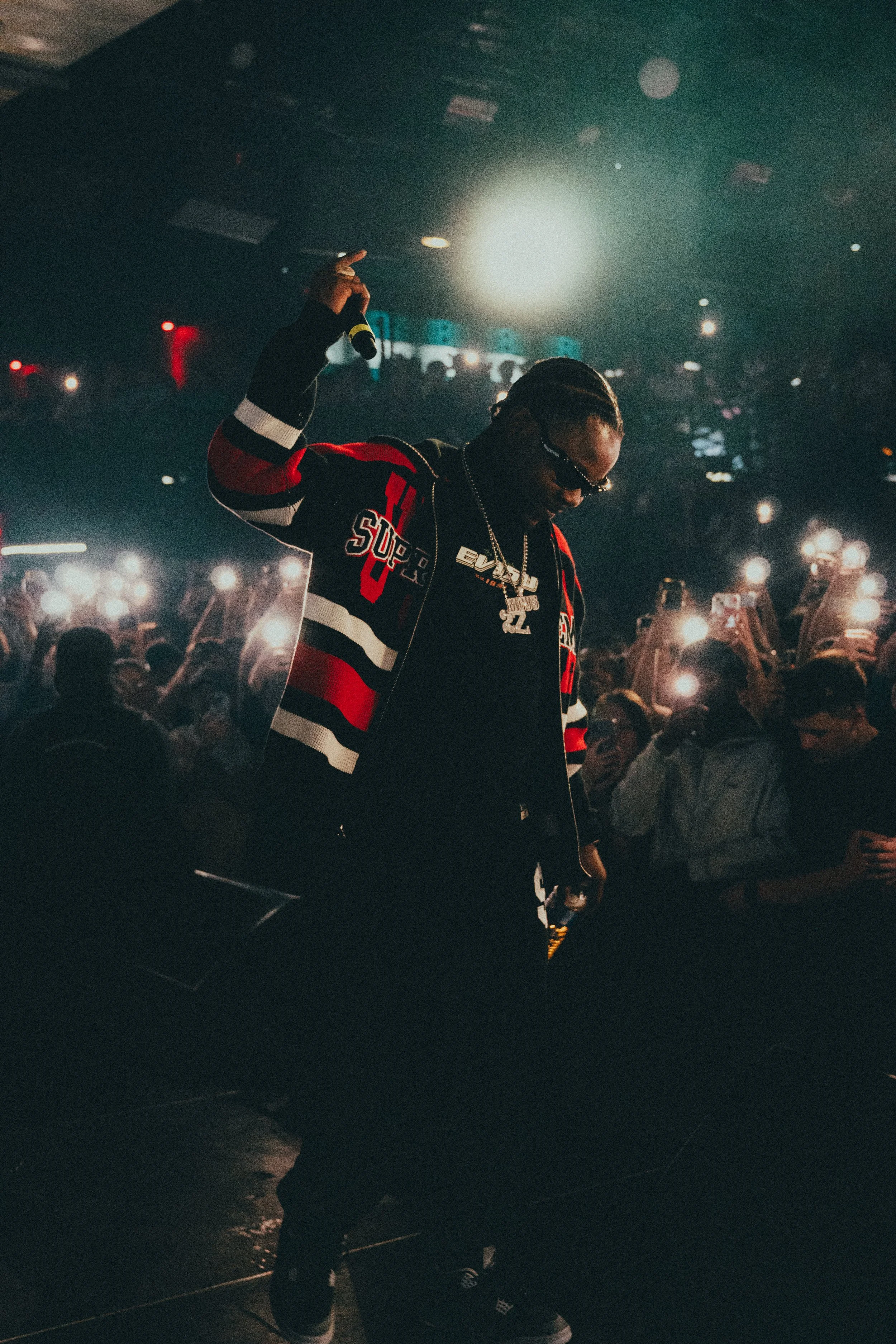 Un artiste en concert shakuant avec un micro, portant des lunettes de soleil et un collier, entouré d'une foule utilisant leur téléphone pour capturer le moment, dans une ambiance sombre et éclairée par des lumières de scène.