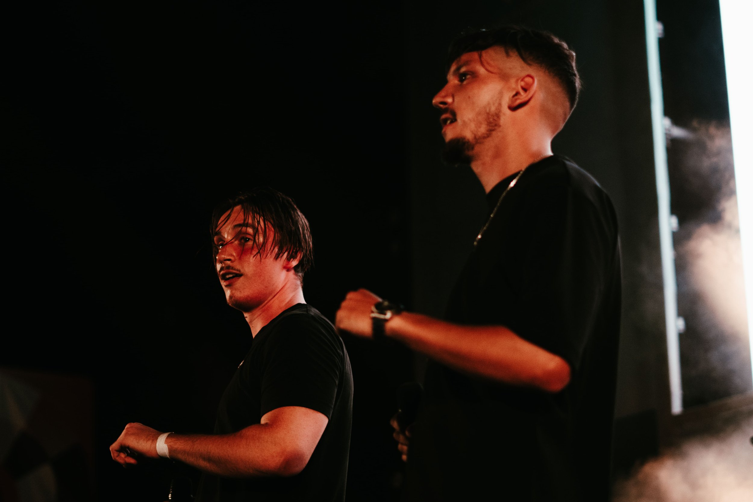 Deux jeunes hommes avec des t-shirts noirs, un aux cheveux longs et mouillés, l'autre coiffé court, se tiennent sur scène dans une ambiance sombre avec un éclairage de scène.»