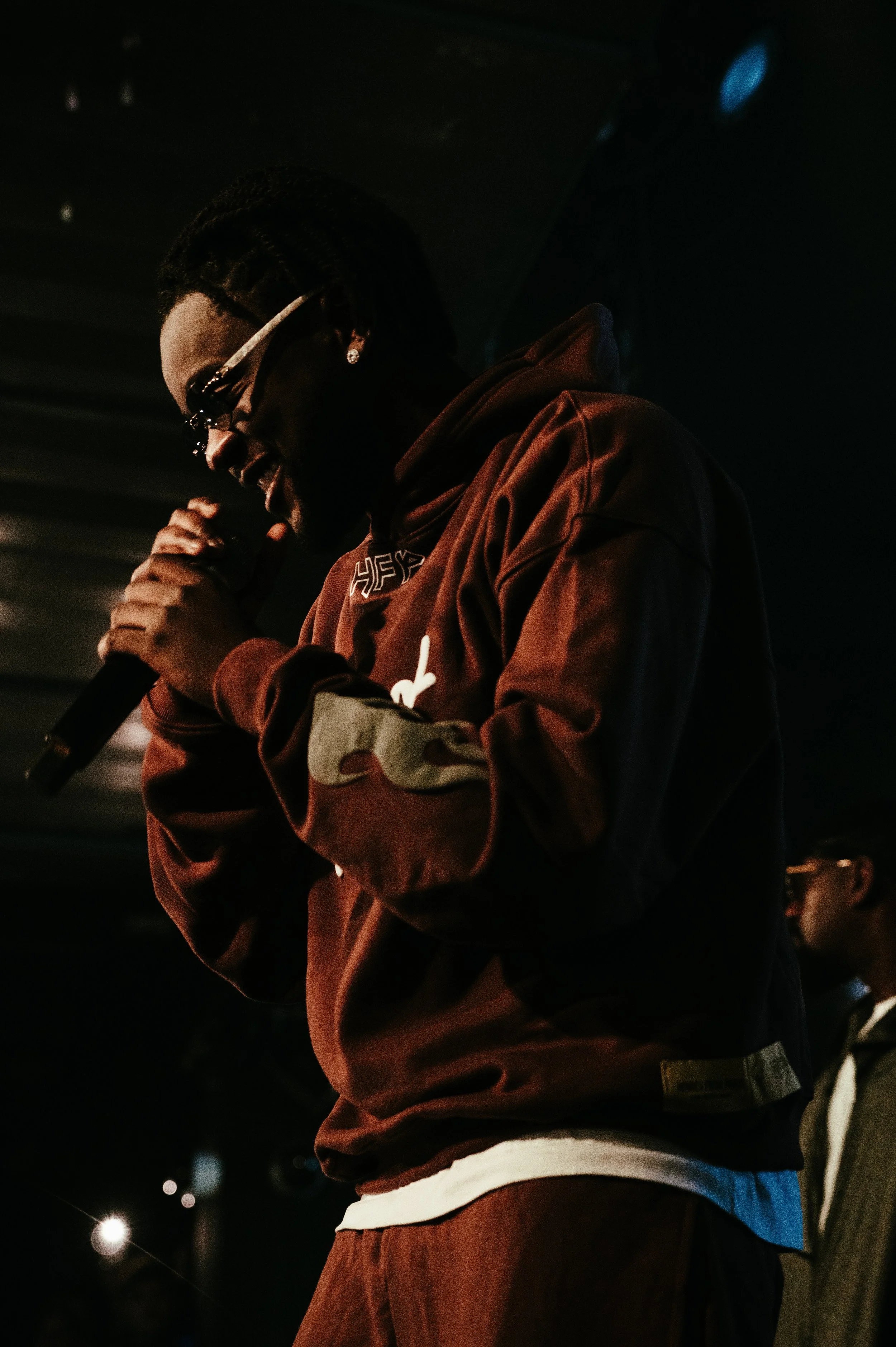 Un homme tenant un microphone, portant une veste marron, des lunettes de soleil et des boucles d'oreilles, parlant ou chantant dans un environnement sombre.