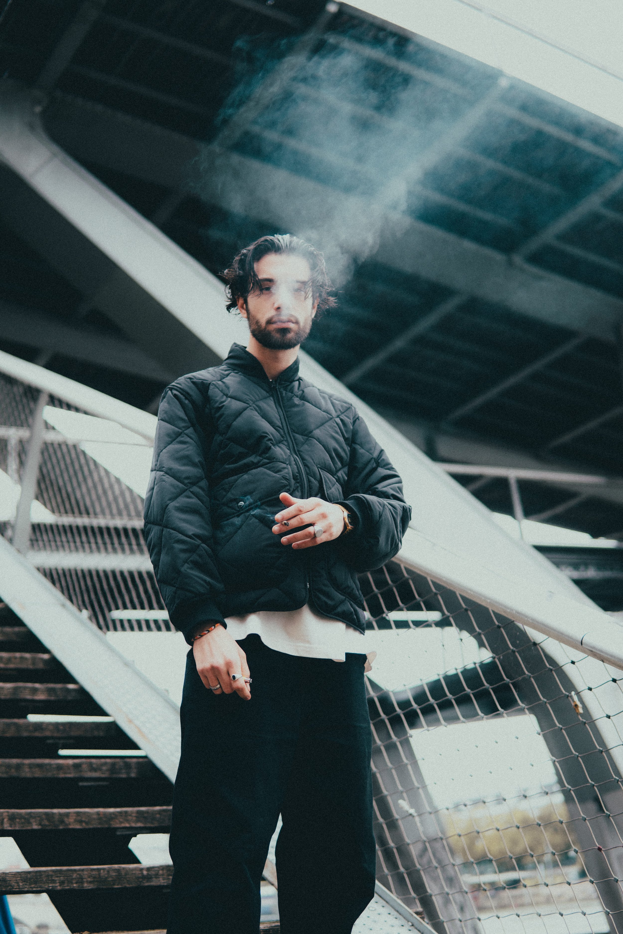 Un homme avec des cheveux noirs, barbe, portant une veste noire, sous un pont ou une structure métallique, avec un escalier en arrière-plan.