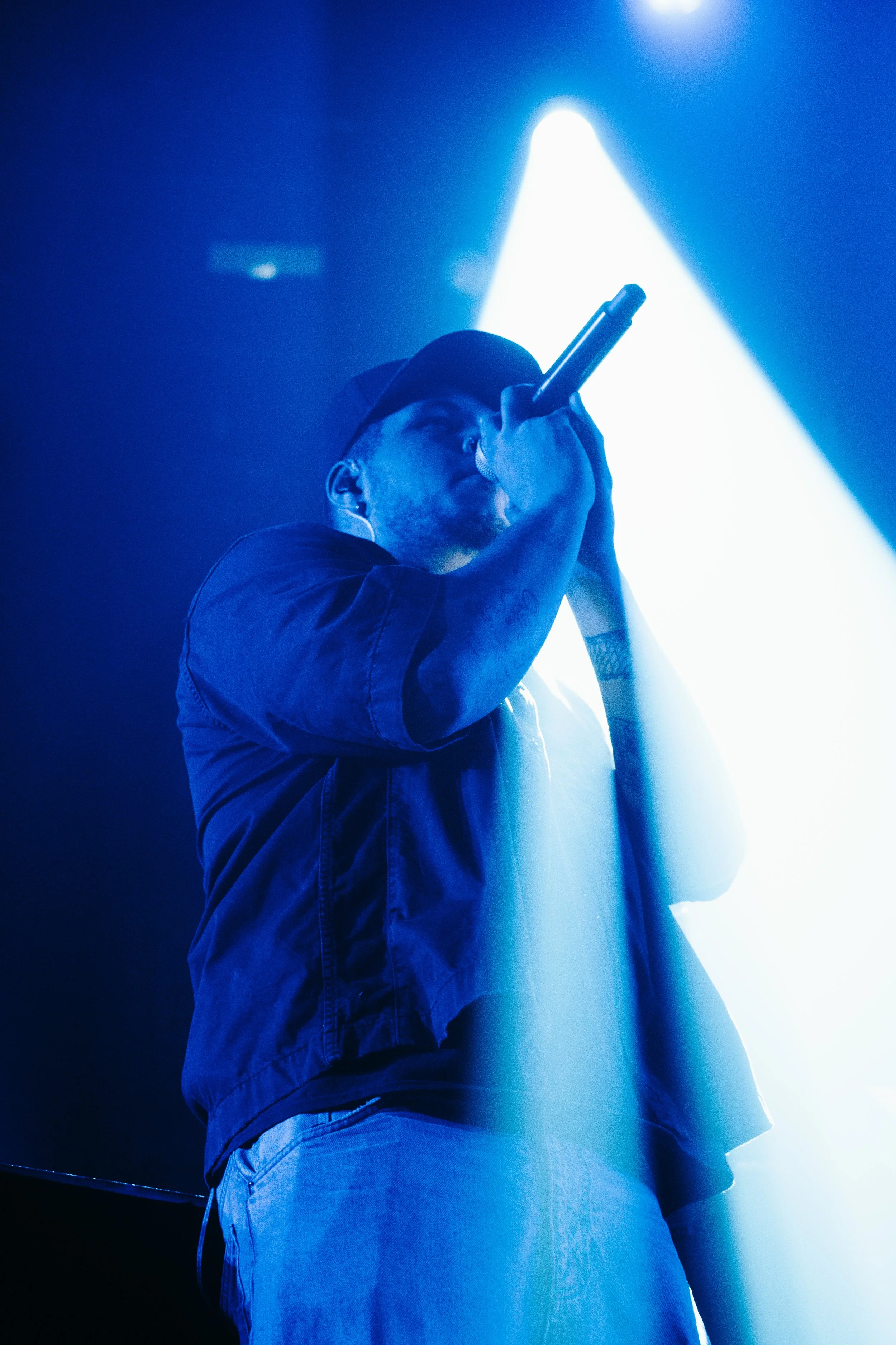 Un homme qui chante ou parle dans un micro lors d'une performance dans un environnement sombre avec un éclairage blanc brillant derrière lui.