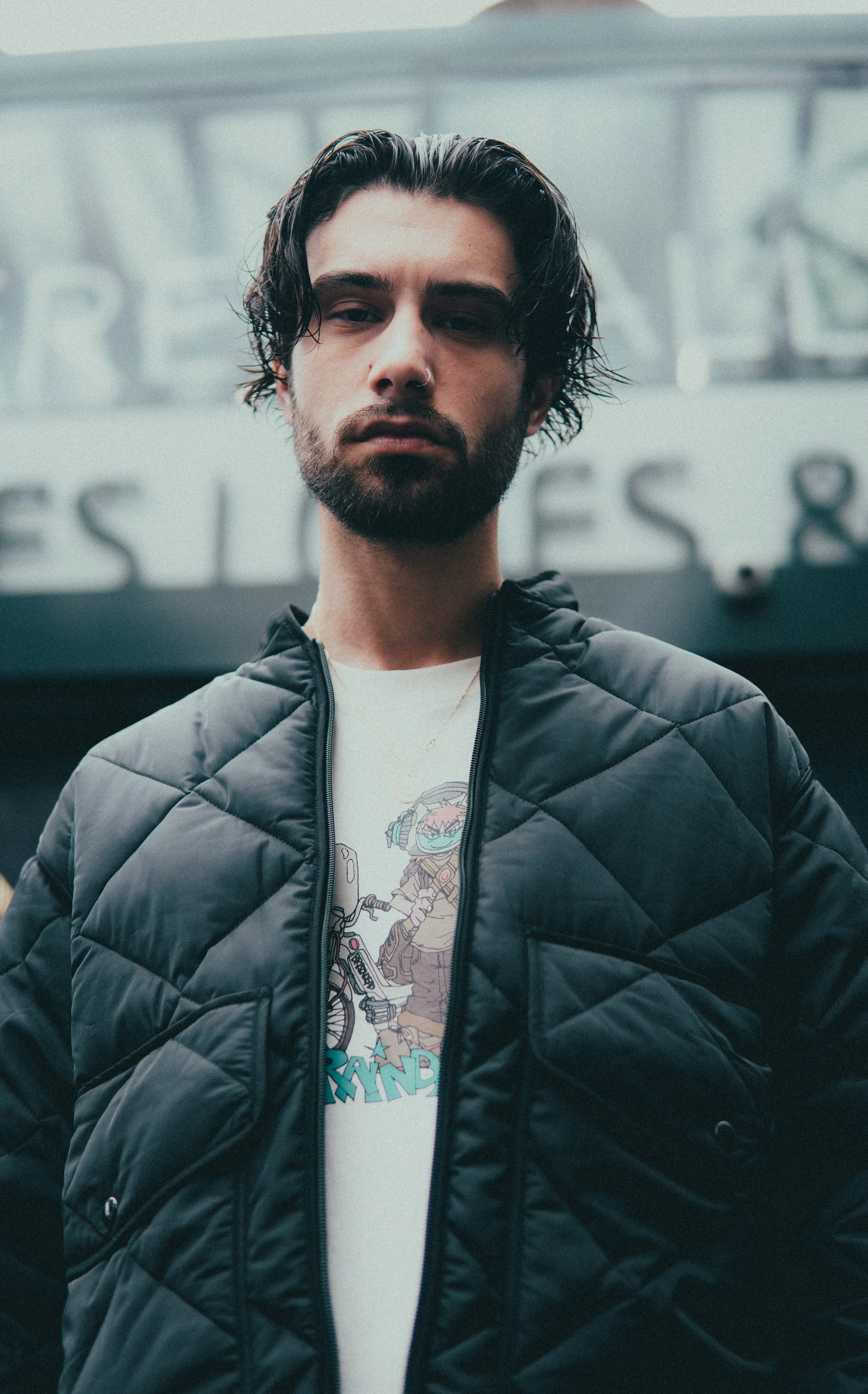 Un jeune homme avec une barbe et des cheveux longs, portant une veste noire en nylon et un t-shirt à motif, dans un environnement urbain.