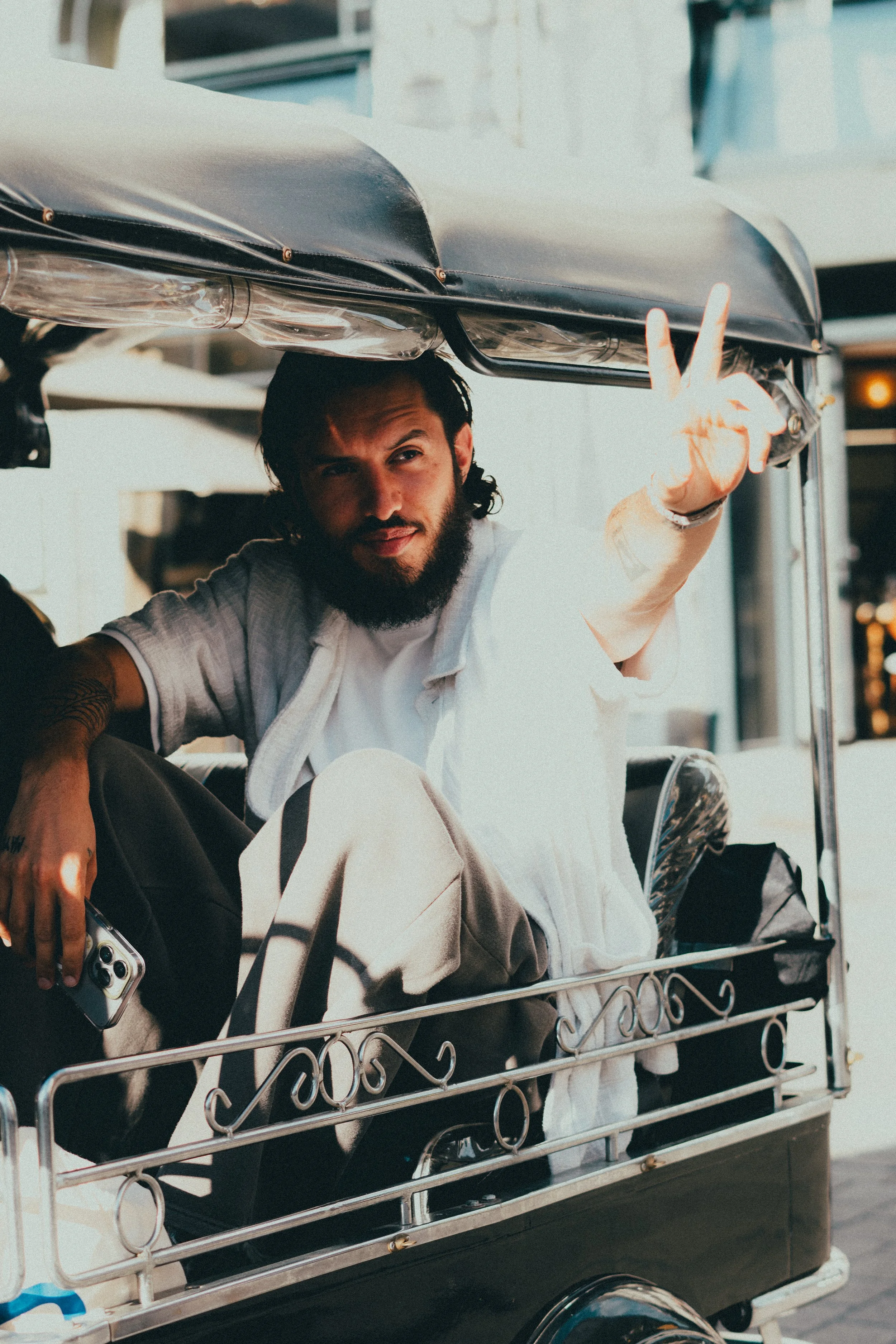 Un homme avec barbe et cheveux foncés, assis dans un tuk-tuk, faisant un signe de paix avec la main droite, tenant un smartphone dans la main gauche, portant une chemise blanche et un pantalon beige, avec un sac à dos noir à côté.