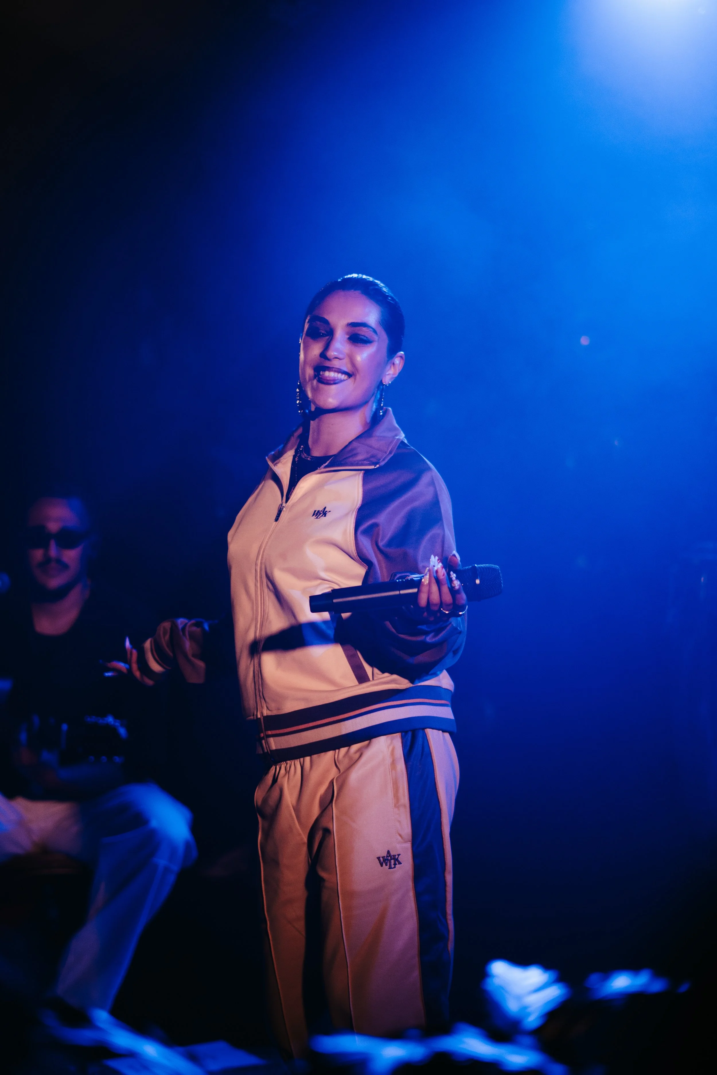Une femme en vêtements de sport colorés, tenant un micro, sur scène avec un fond lumineux bleu, souriante et en train de perform