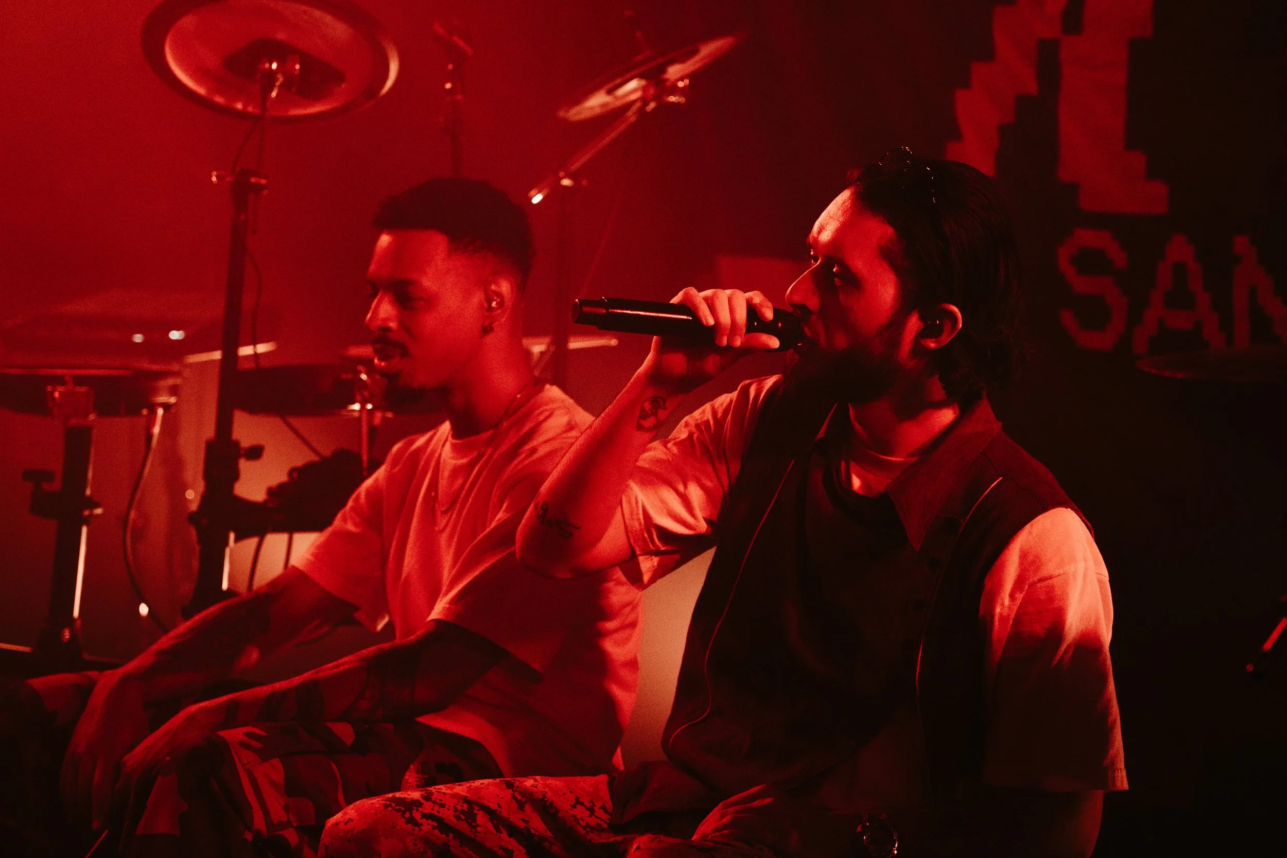 Deux hommes assis sur scène sous un éclairage rouge, avec un humain tenant un microphone.
