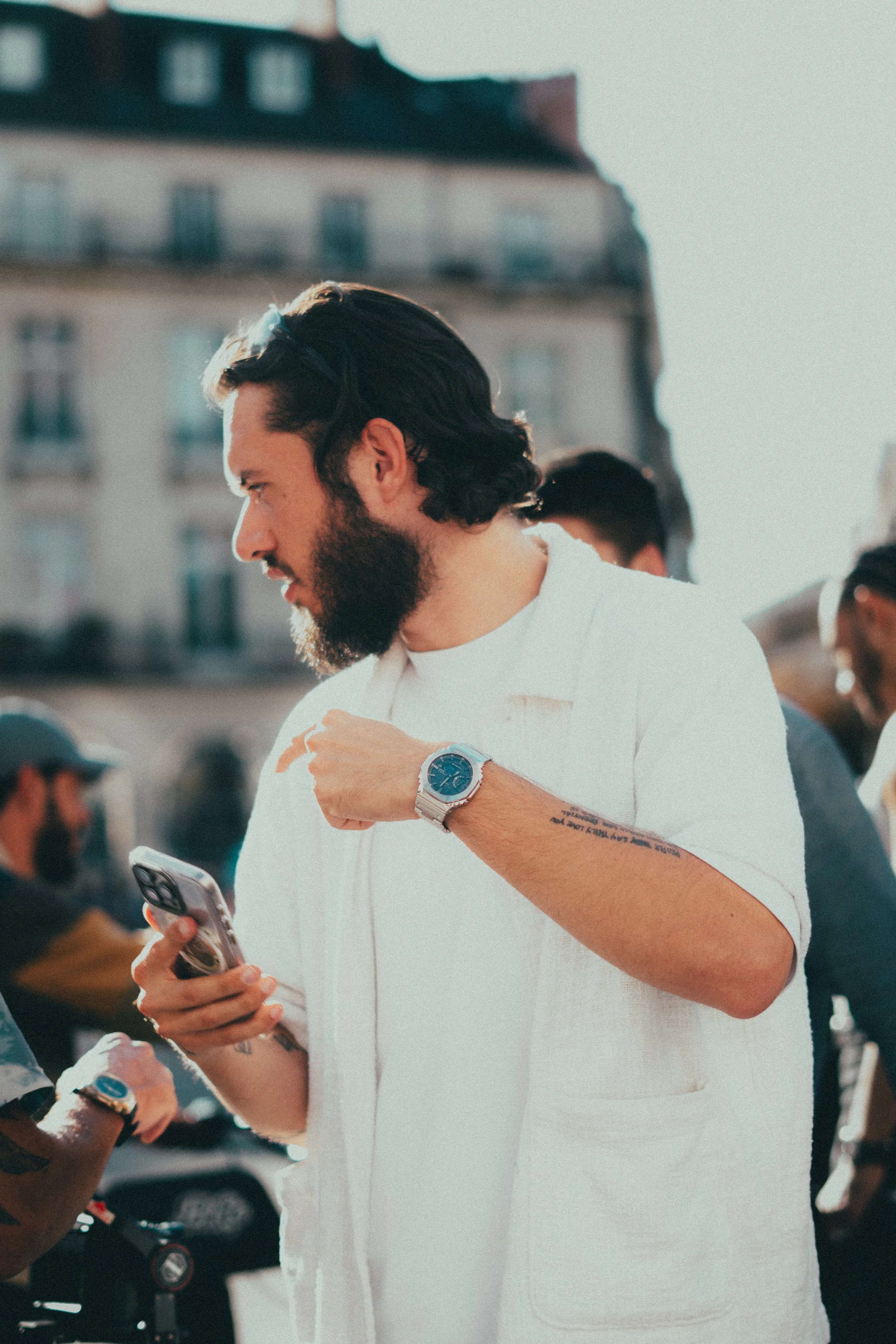 Un homme à barbe, portant une montre en métal, regarde son téléphone cellulaire en plein air, avec un bâtiment résidentiel en arrière-plan.