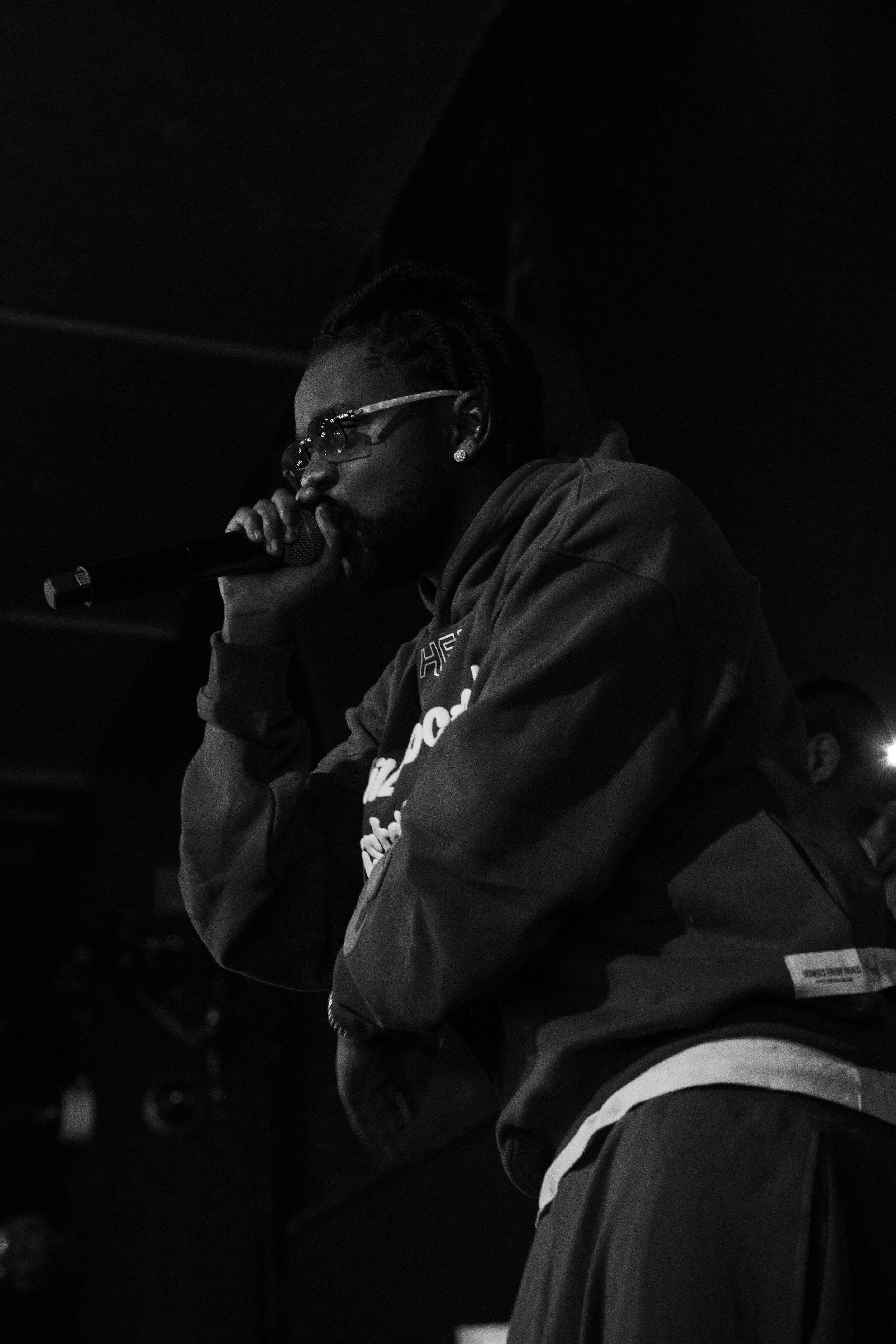 Un homme portant des lunettes et une veste, tenant un microphone près de la bouche, en train de chanter ou de parler dans un espace sombre.