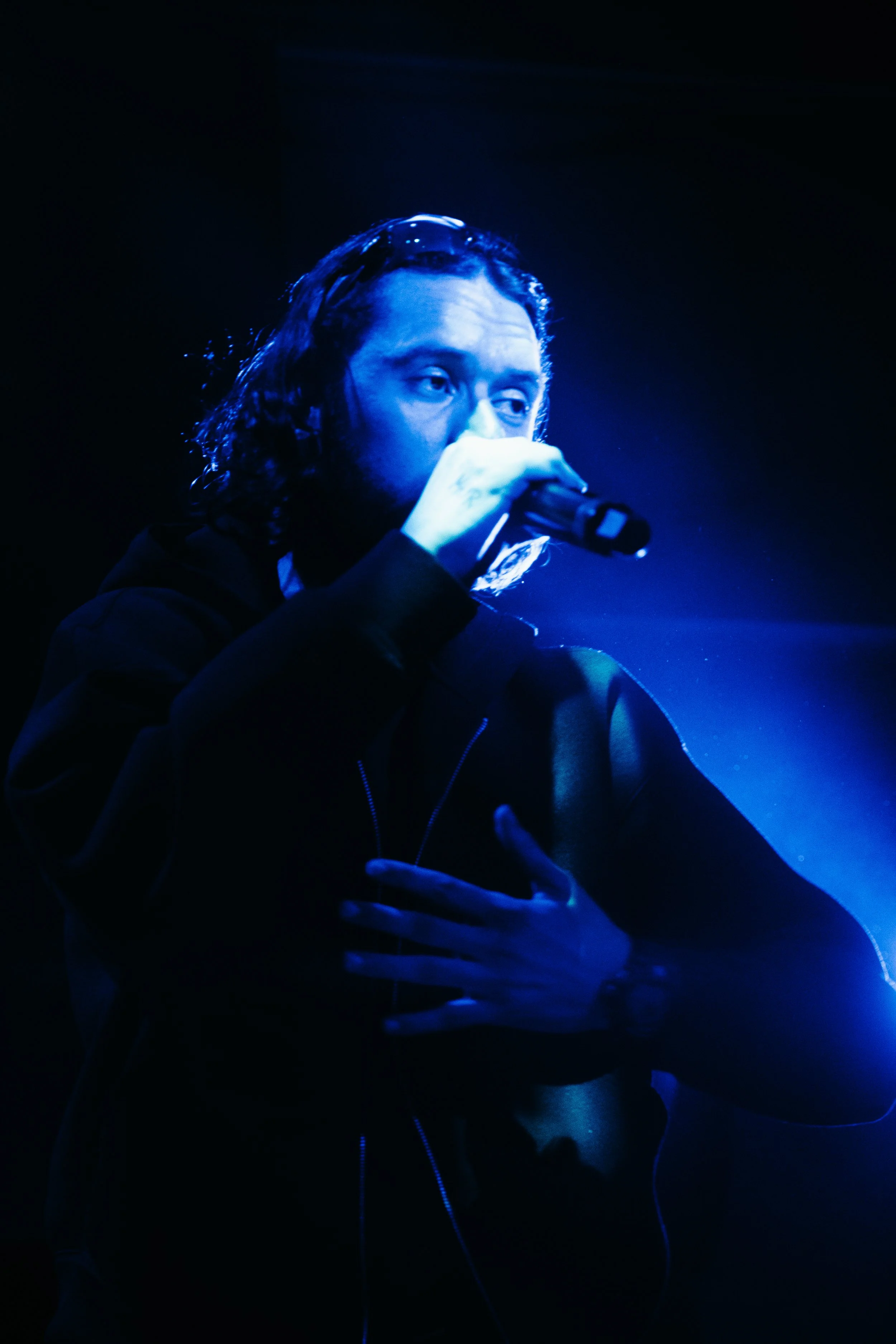 Un homme avec des cheveux longs chante ou parle dans un microphone, éclairé en bleu dans une scène sombre.