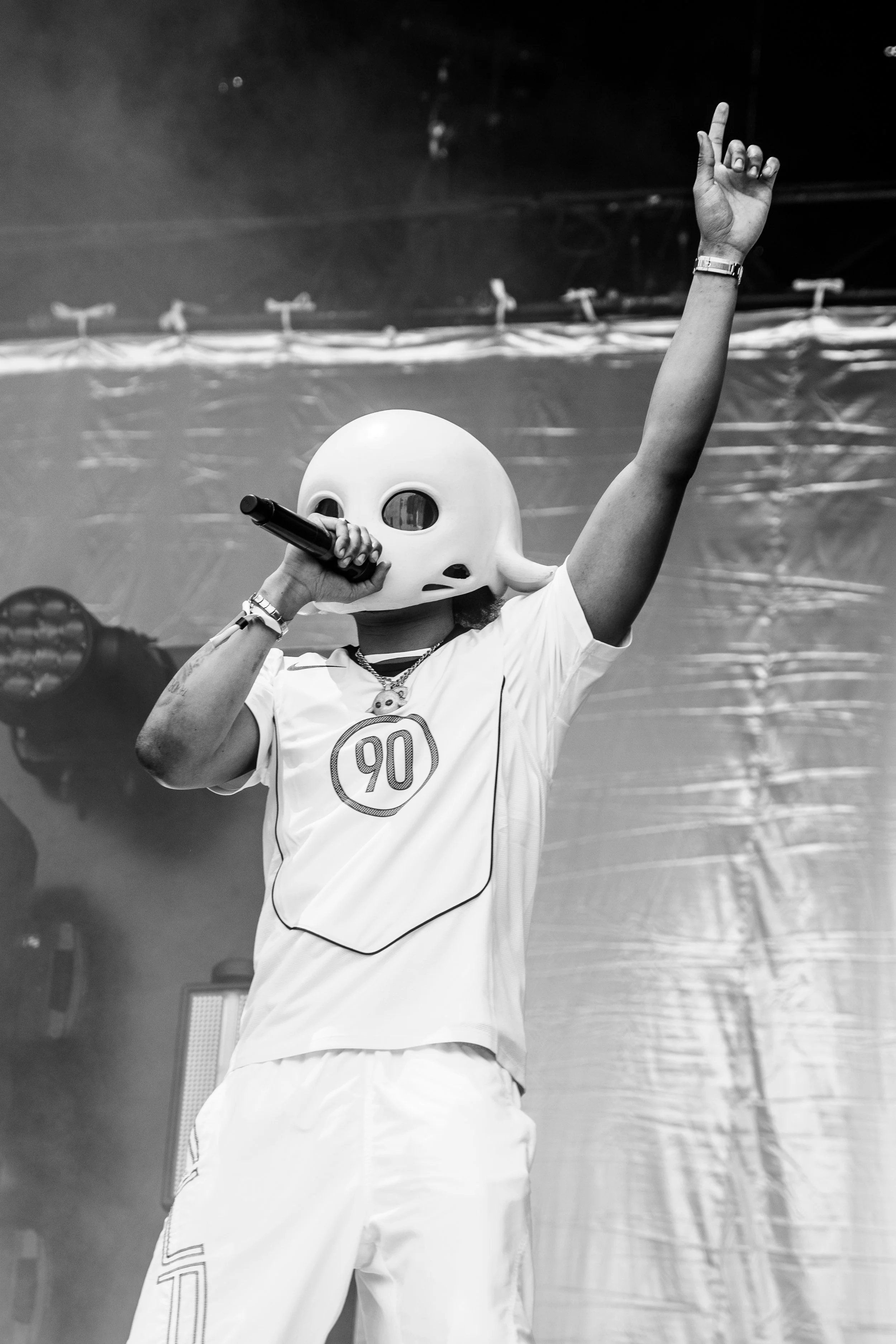 Une personne porte un masque de tête de fantôme blanc, tient un microphone dans une main et lève l'autre main avec le index pointé vers le haut. La personne porte un t-shirt blanc avec le numéro 90. L'arrière-plan est sombre avec un éclairage de scèn