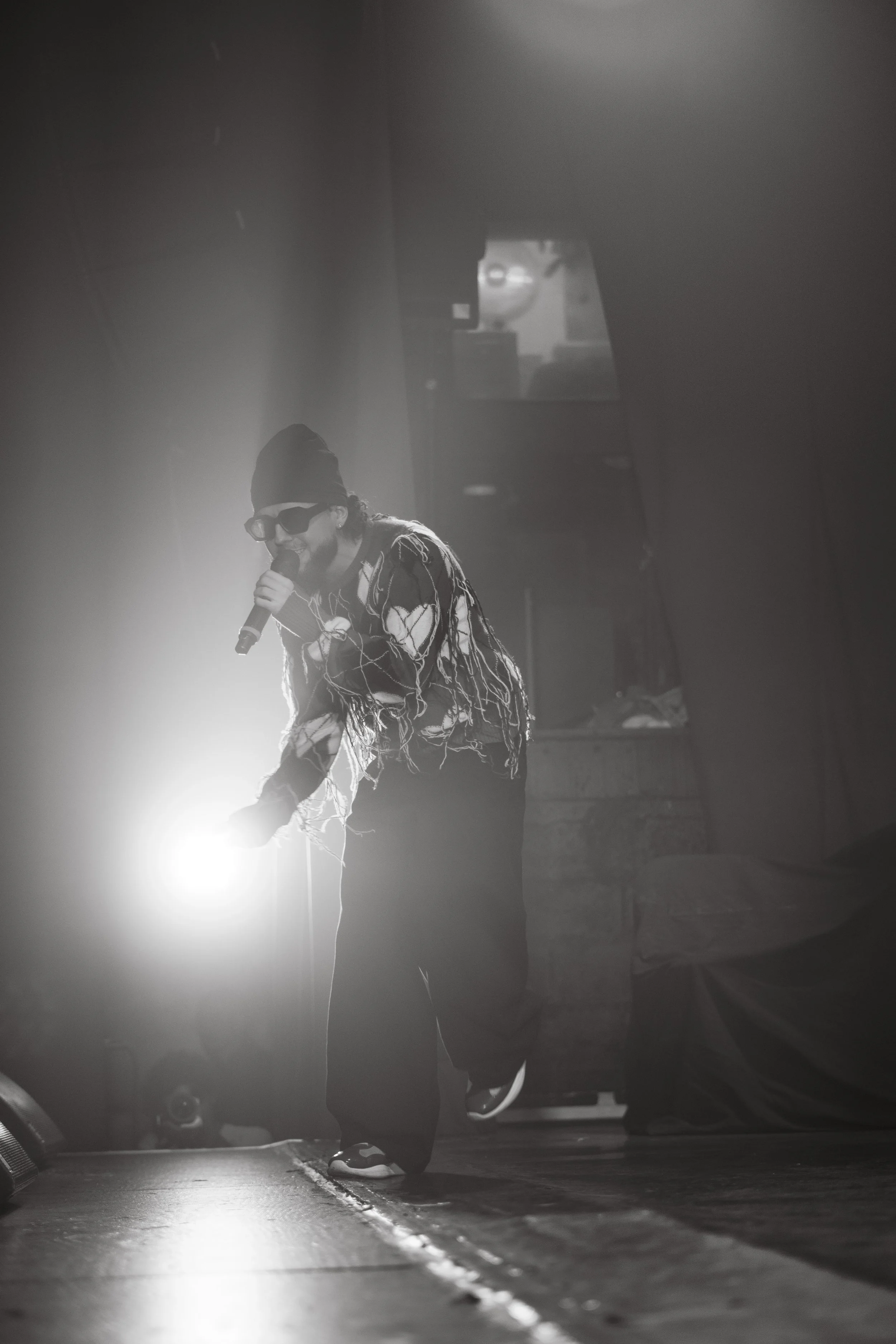 Un artiste avec des lunettes noires et un bonnet, tenant un microphone, en train de performer sur scène avec un éclairage intense. En arrière-plan, une scène sombre avec une fenêtre et des équipements.