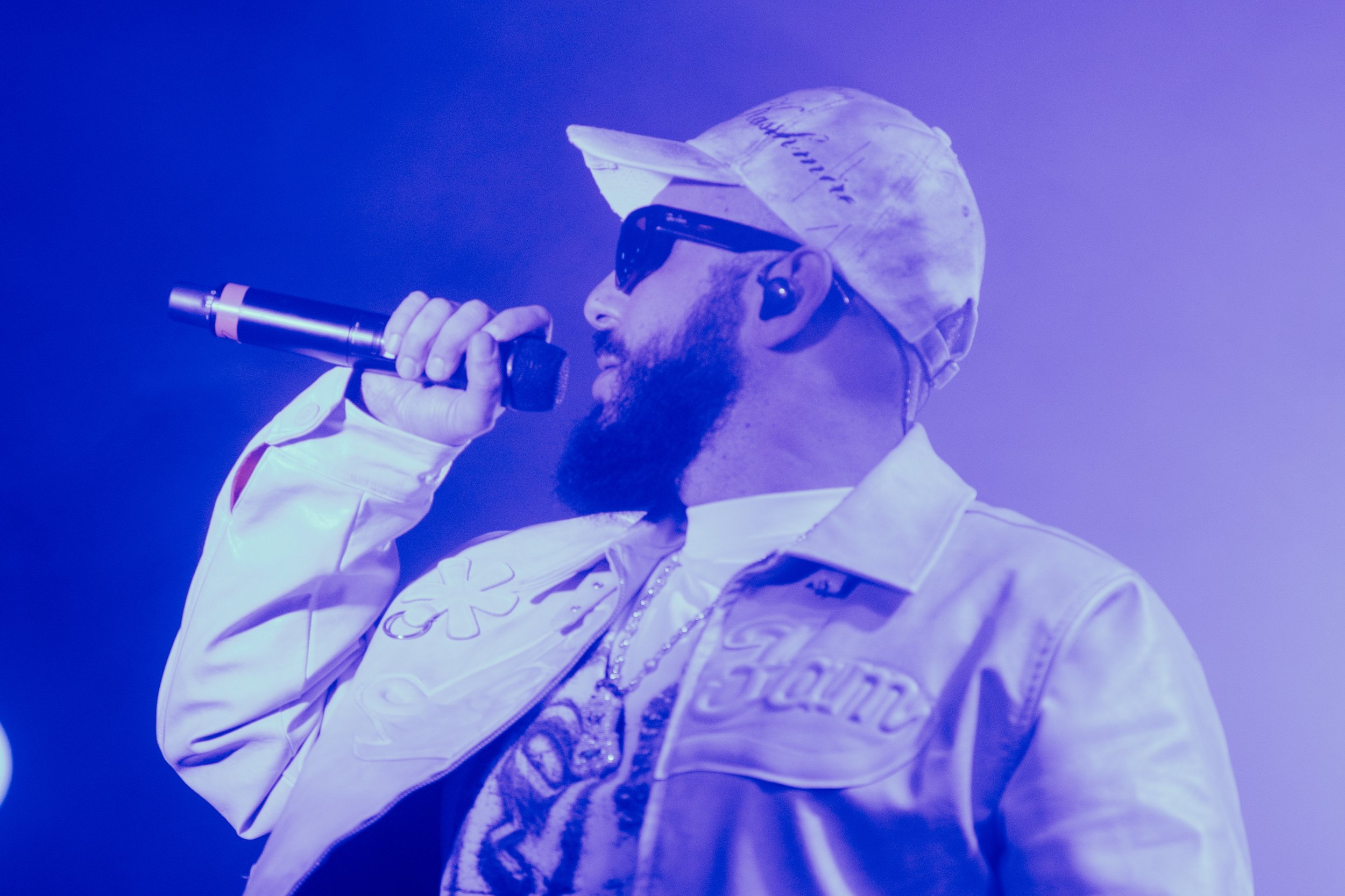 Un homme avec une barbe, portant une casquette, des lunettes de soleil, et une veste blanche avec des motifs, tient un microphone et chante ou parle. L'image est en teintes de violet et de bleu.