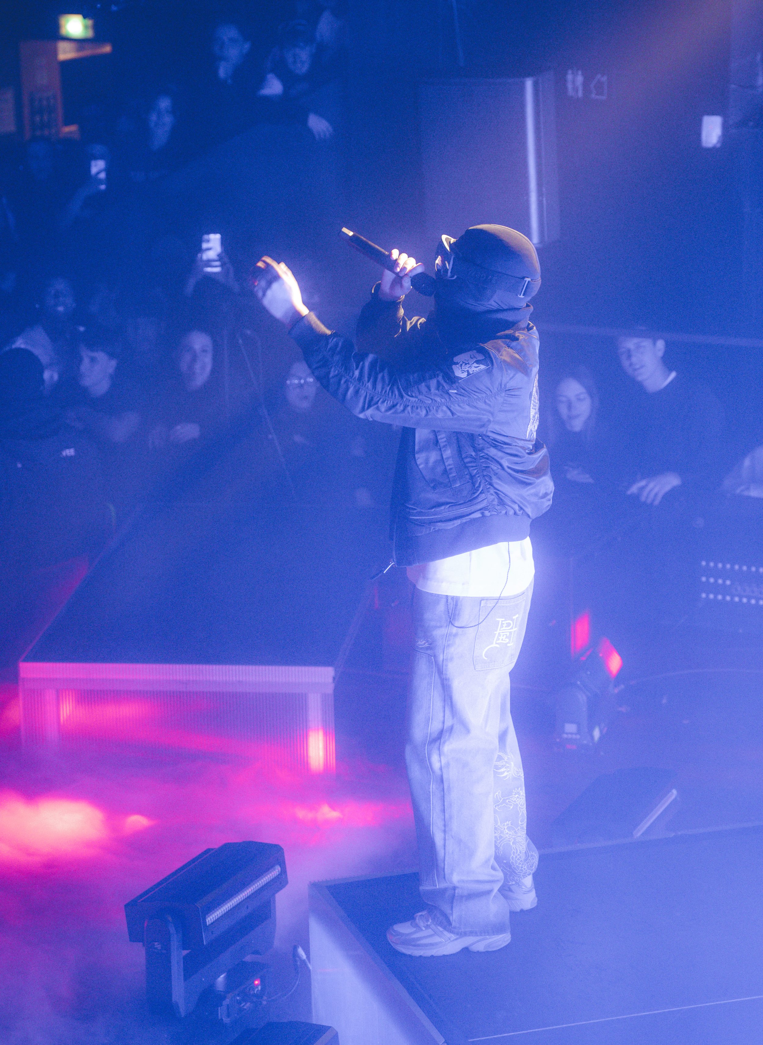 Un artiste en train d'performer avec un micro sur scène, entouré d'un public dans une ambiance sombre avec des lumières violettes et roses.