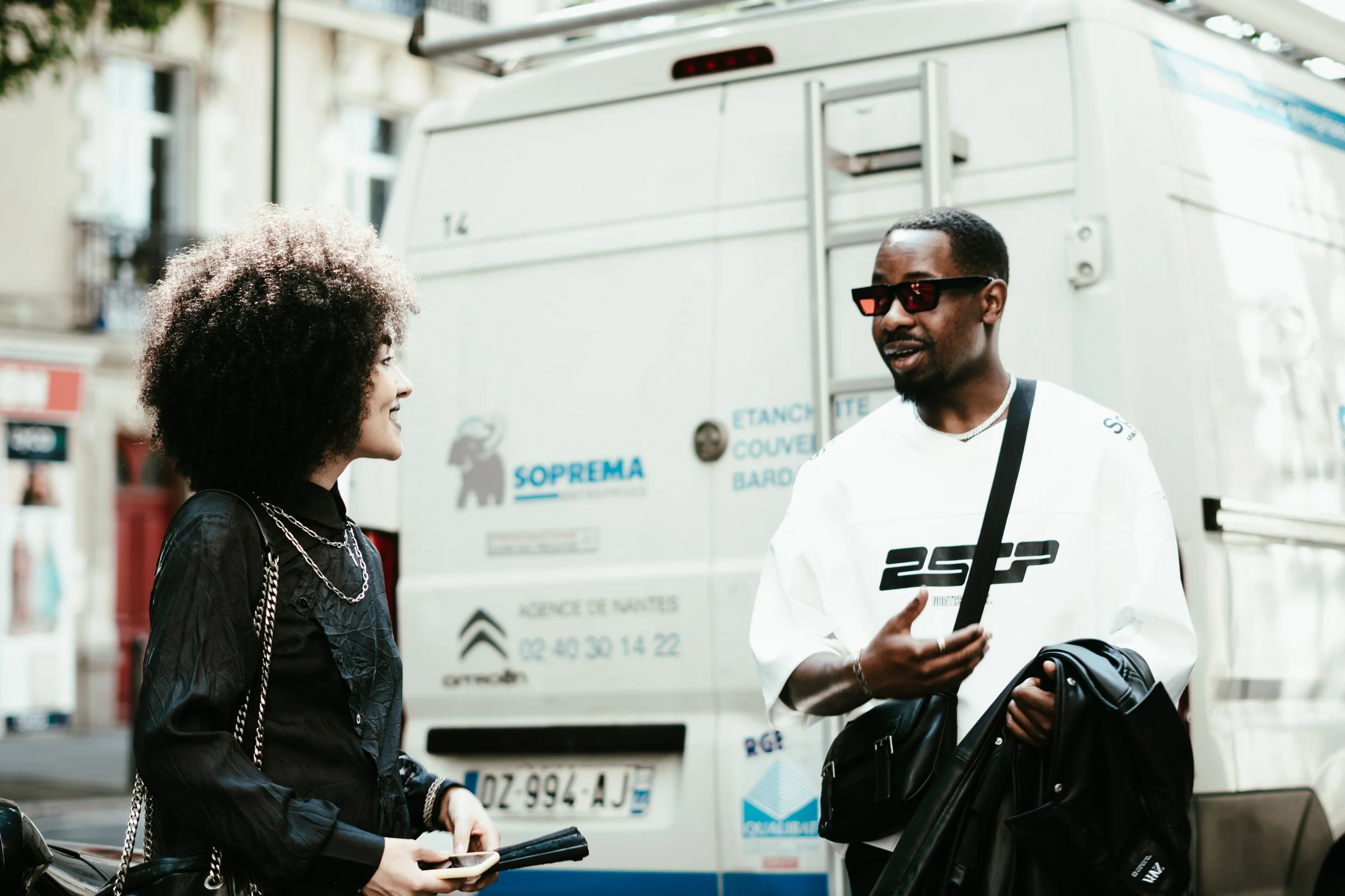 Deux personnes discutent dans la rue. L'homme porte des lunettes de soleil, un sweat blanc et tient un sac noir, la femme a des cheveux bouclés, portait une veste noire avec des chaînes, et tient un téléphone portable.