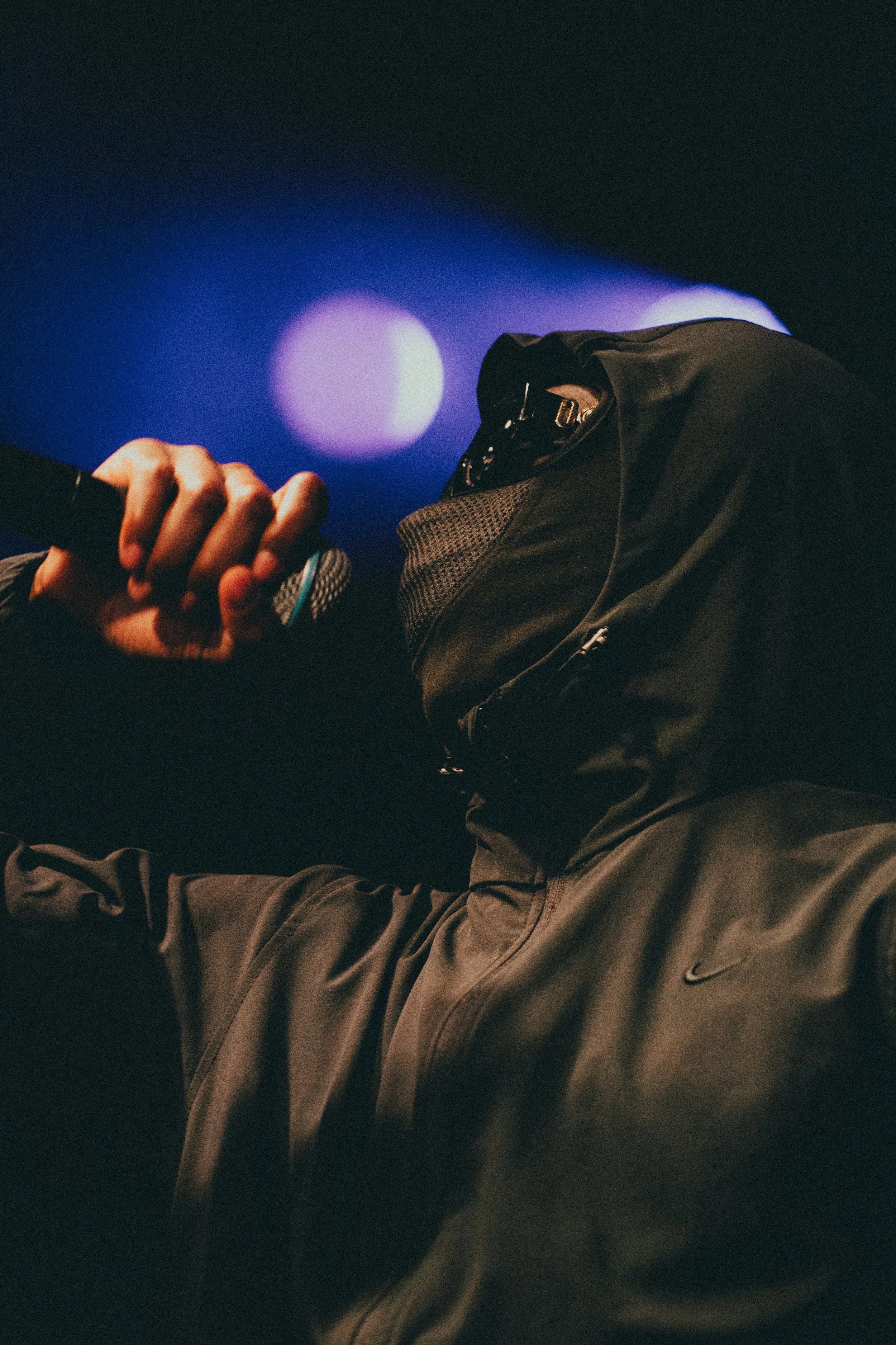 Une personne portant un sweat à capuche noire et un masque de ski tient un microphone lors d'une performance scénique, avec des lumières colorées en arrière-plan.