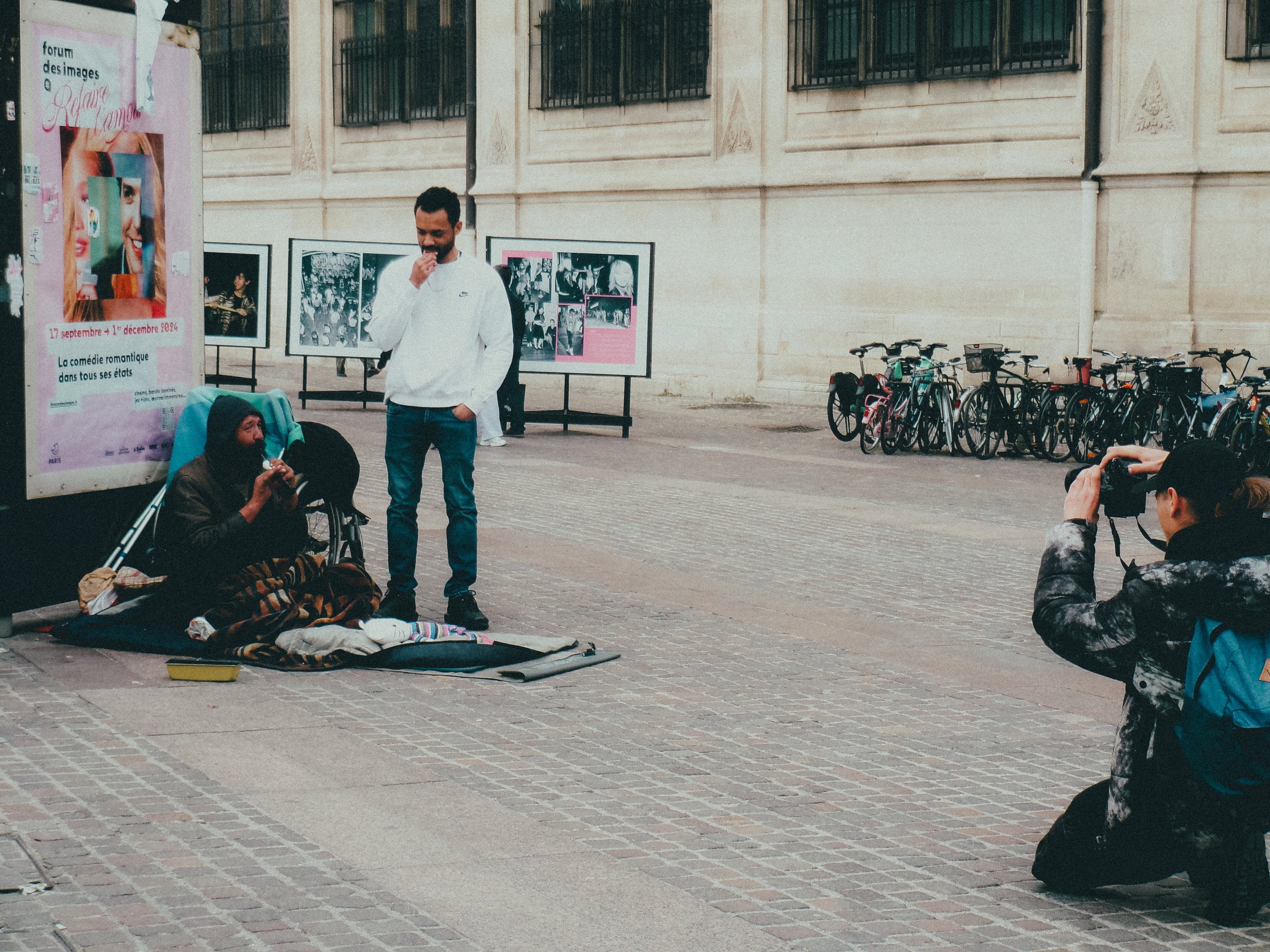 Un homme sans-abri assis sur un matelas, discutant avec un jeune homme debout à côté de lui, tandis qu'une femme prend une photo, sur une rue pavée. Des vélos sont stationnés en arrière-plan, et une affiche publicitaire est visible sur le côté gauche