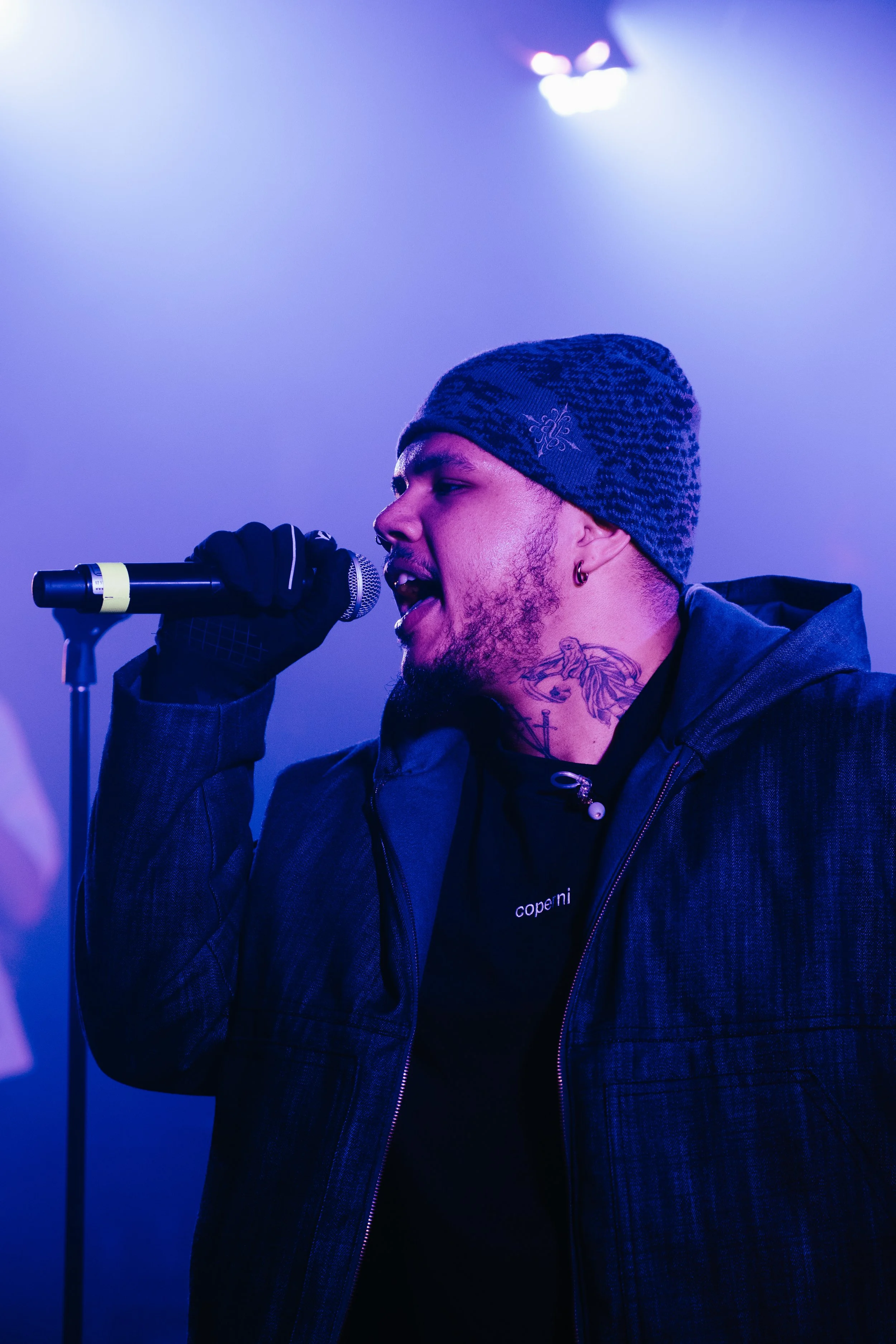 Un homme chantant dans un micro, portant un bonnet et une veste, avec des tatouages visibles sur le cou, sous un éclairage violet.