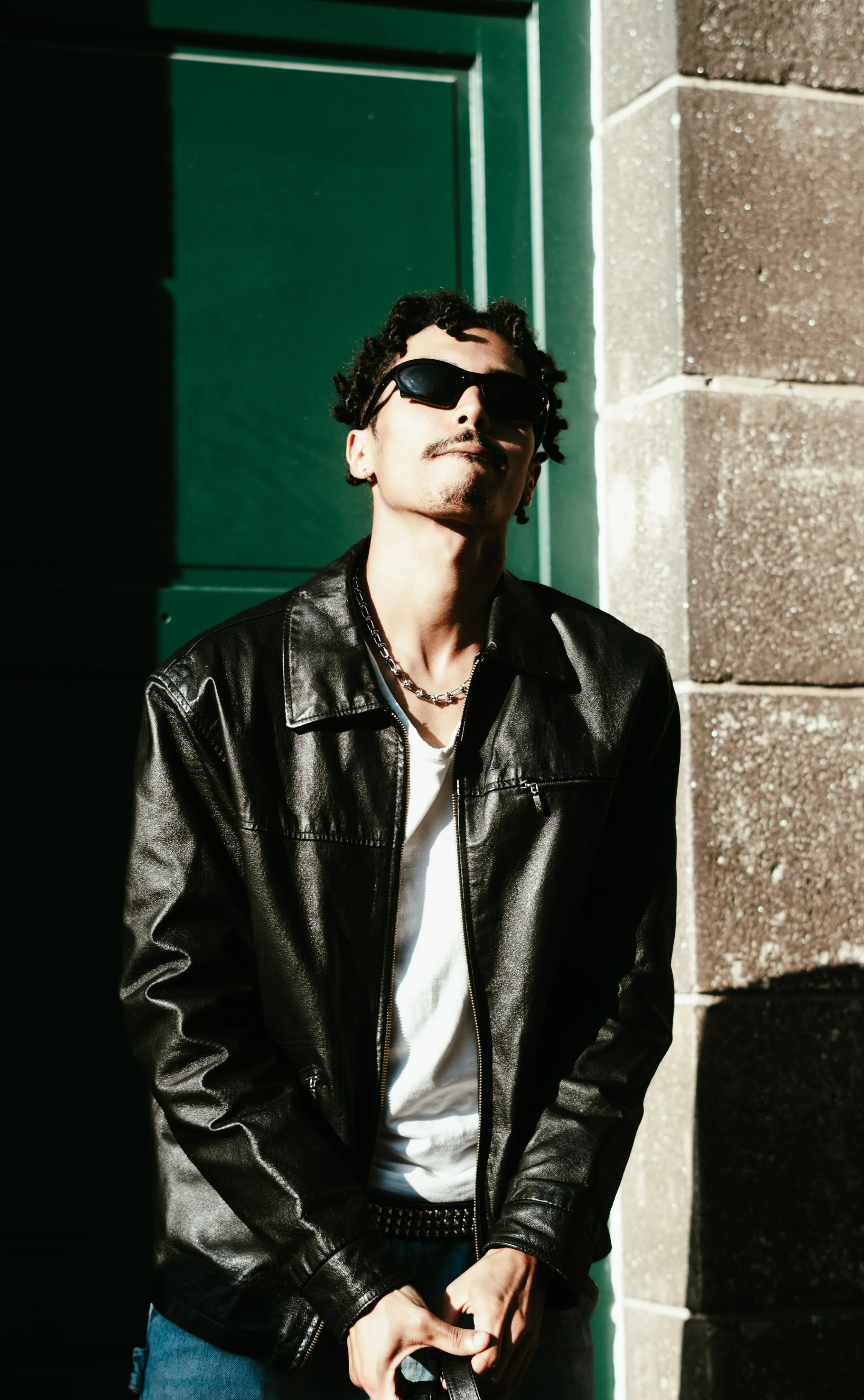 Jeune homme portant des lunettes de soleil, une veste en cuir noir, un t-shirt blanc, un collier en chaîne, posant à côté d'un mur en pierre et d'une porte verte dans une lumière solaire