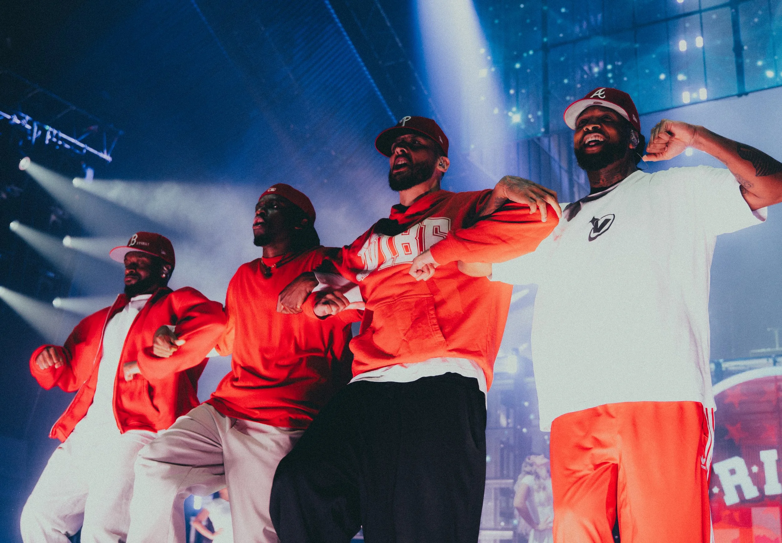 Groupe de quatre hommes lors d'un événement, portant des vêtements de sport aux couleurs rouge, blanc et orange, avec des casquettes. Ils semblent célébrer ou se préparer à une performance sur scène, avec un éclairage de scène sous un plafond moderne