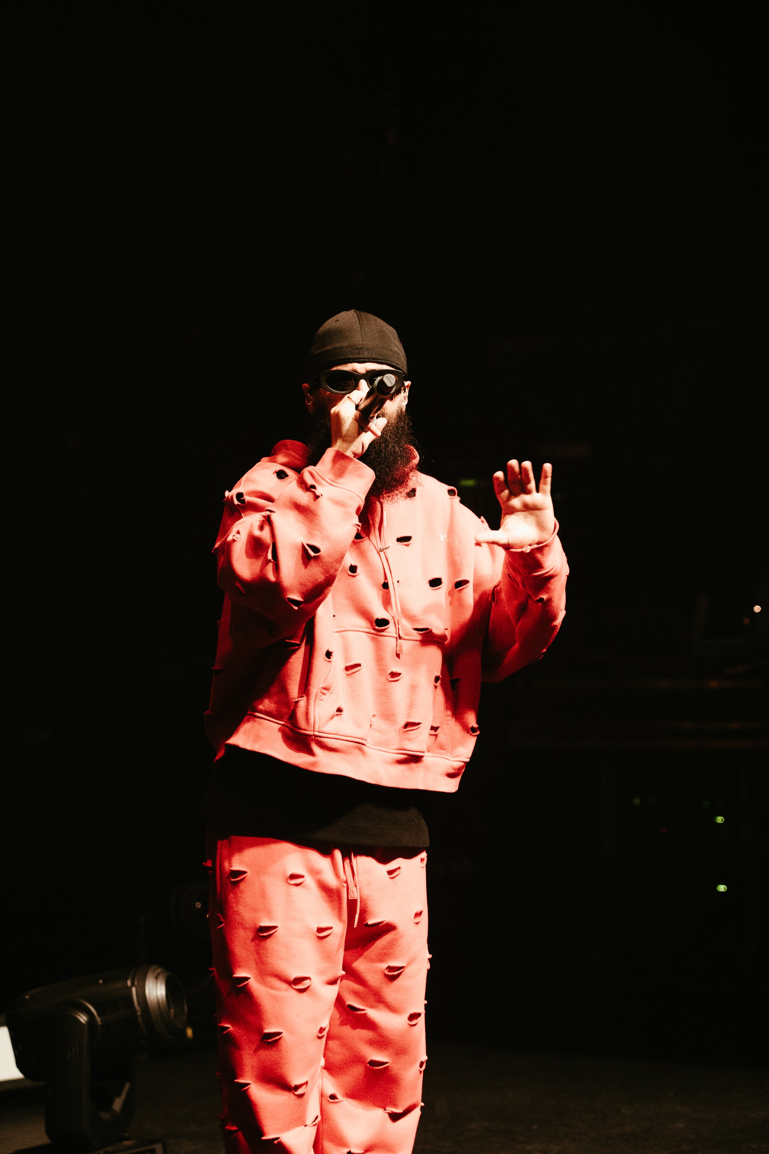Un homme avec une barbe portant des lunettes de soleil, un bonnet noir, un sweat-shirt et un pantalon rose avec des trous, tenant un microphone sur scène.