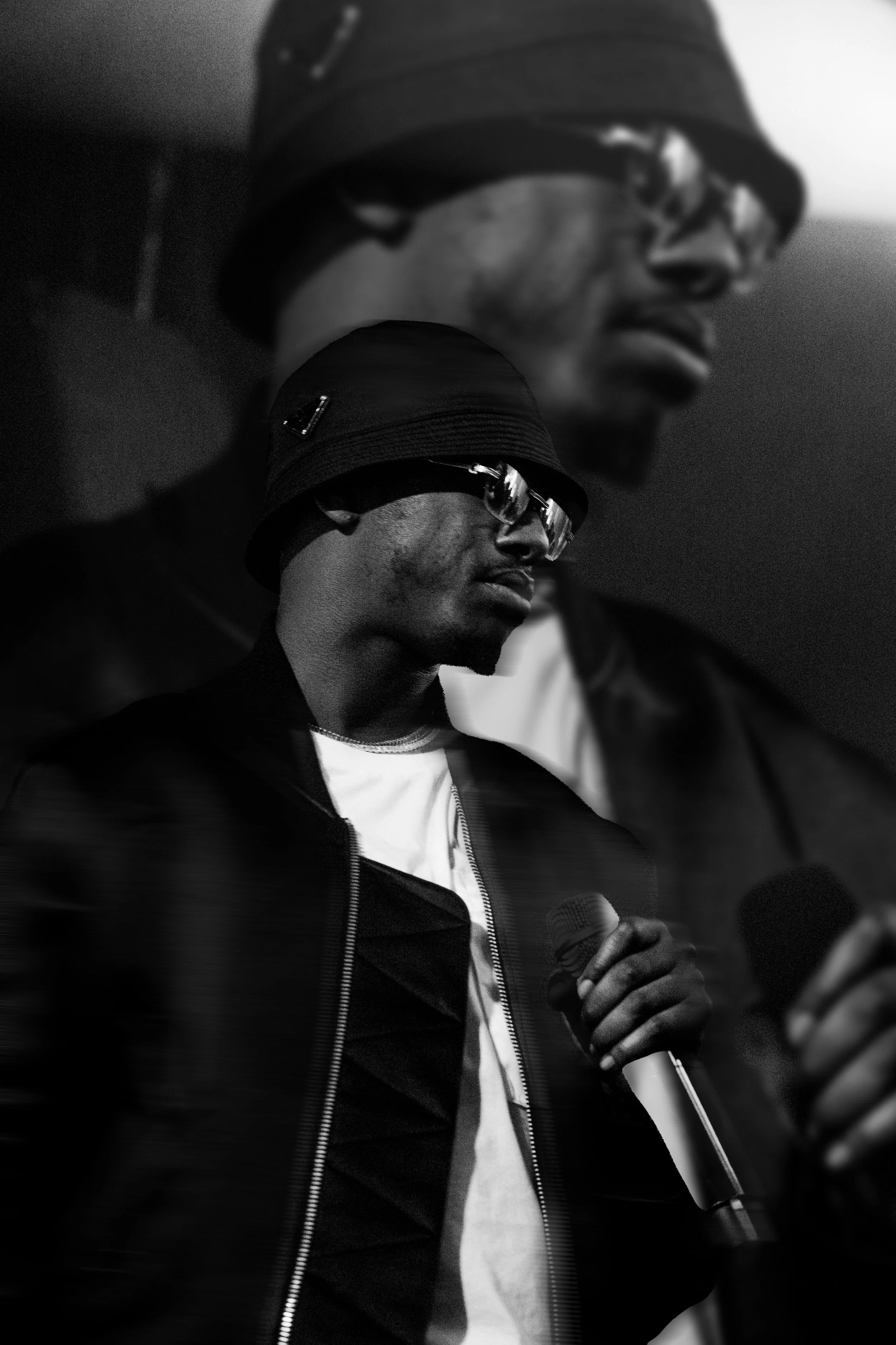 Un homme portant un chapeau noir, des lunettes de soleil, un t-shirt blanc et une veste noire, tenant un microphone en main, dans un contexte sombre.