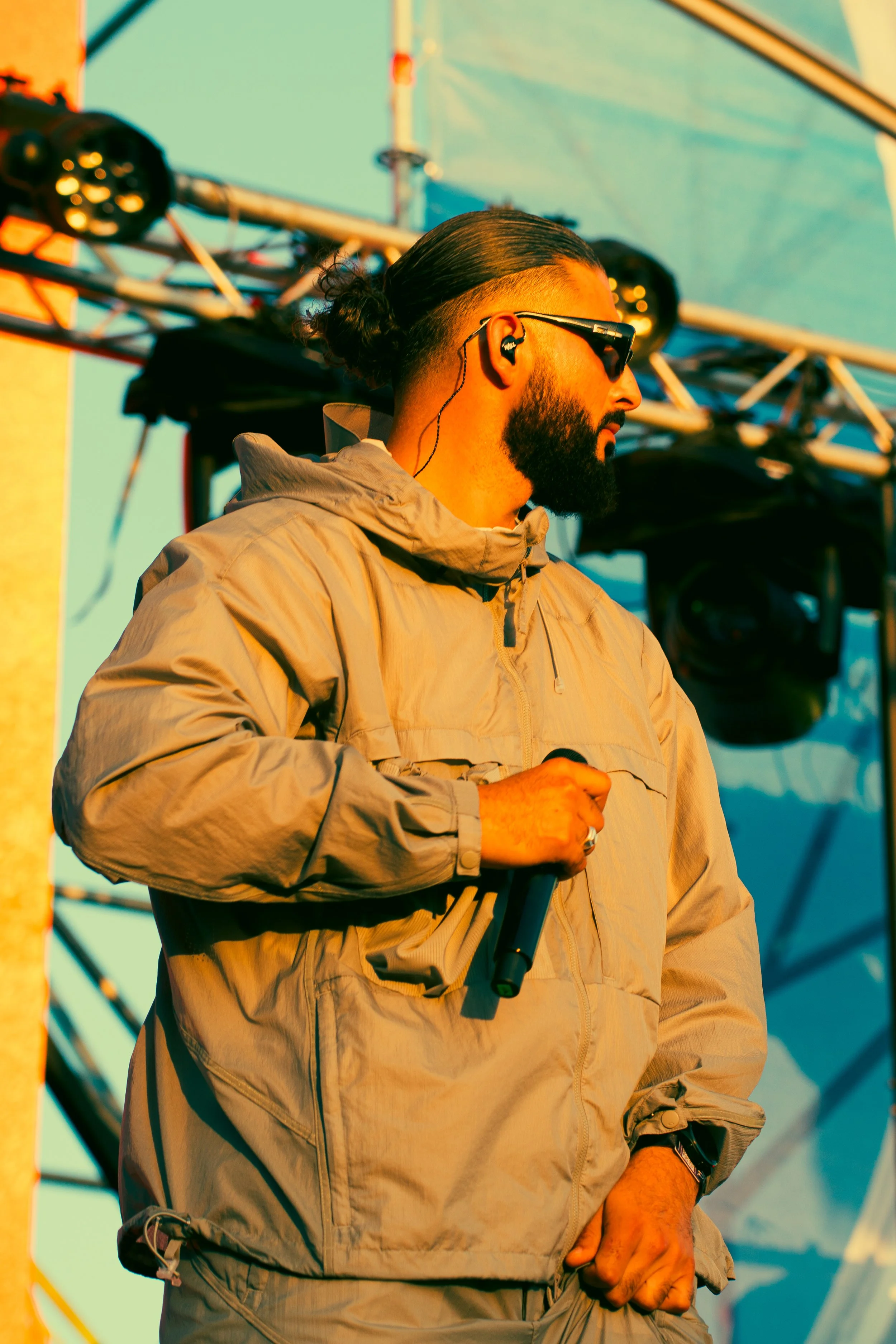 Un homme portant des lunettes de soleil, une veste beige, tenant un microphone et portant des écouteurs, se tient devant une scène avec une structure métallique et des projecteurs, lors d'un spectacle en plein air.