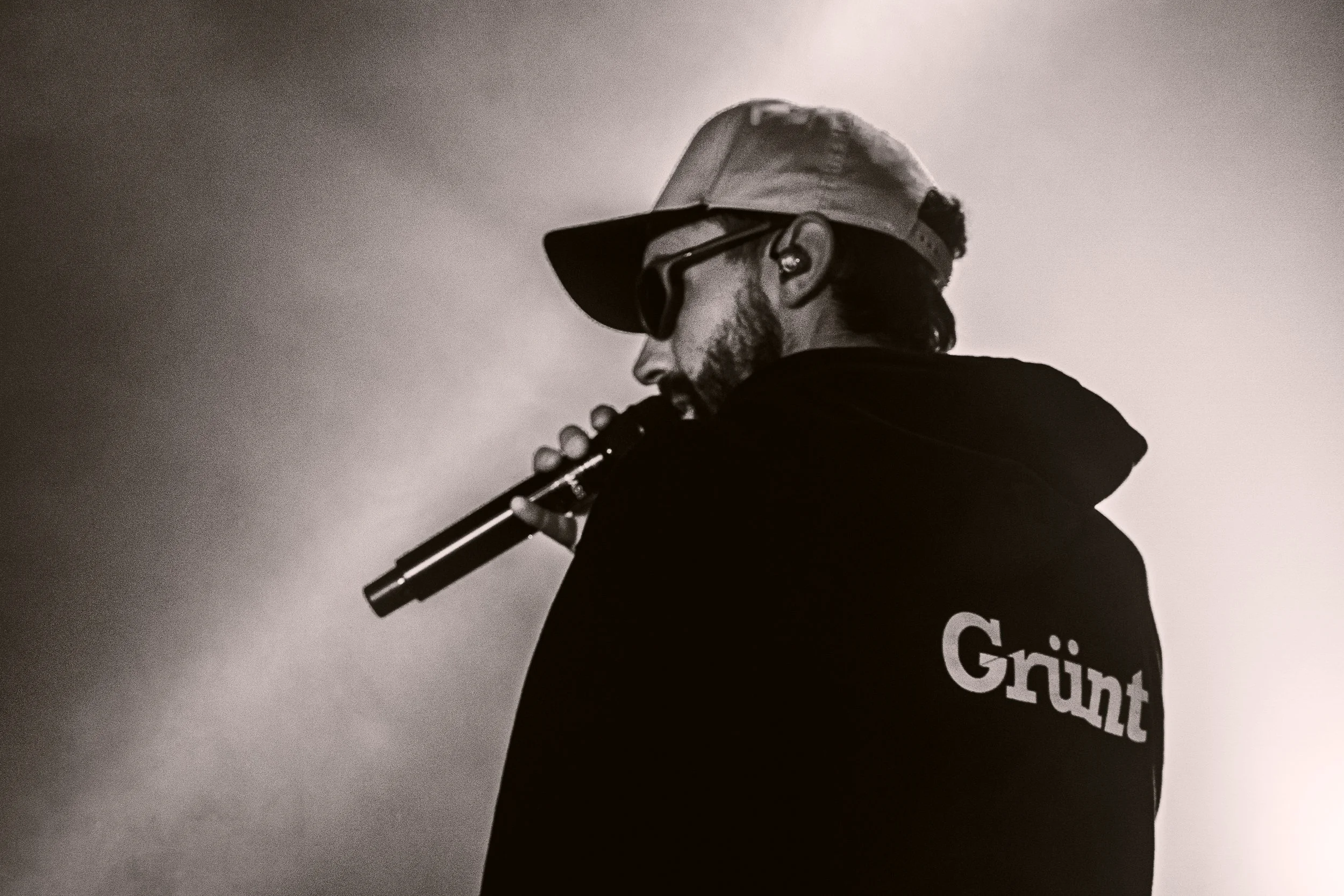 Un homme portant une casquette et des lunettes de soleil joue de la flûte, portant un sweat-shirt avec le mot 'Grünt' à l'arrière, dans une scène en noir et blanc.