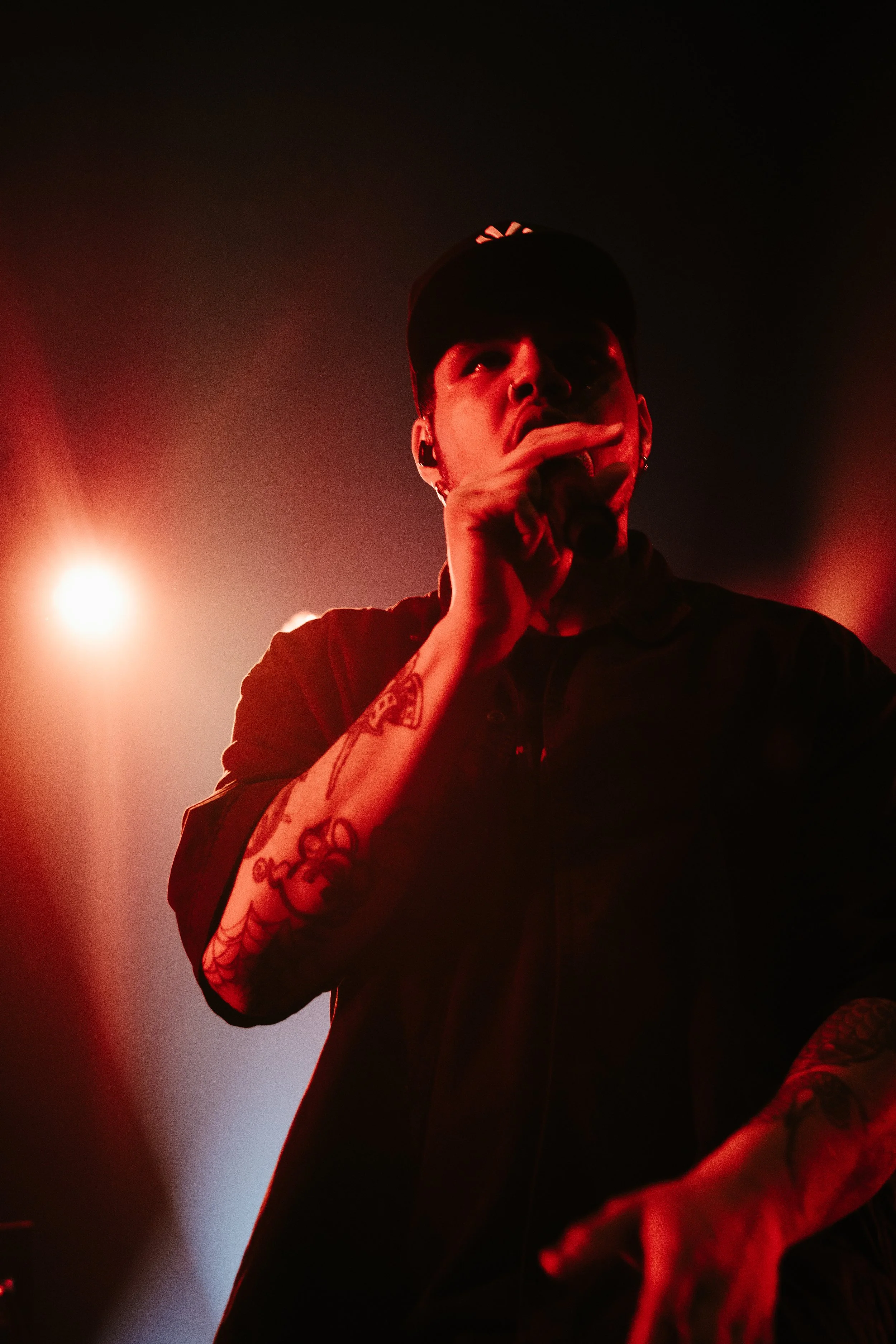 Un homme chante ou parle dans un microphone lors d'un concert, portant une casquette noire et un tatouage visible sur le bras, avec des lumières de scène rouges et blanches en arrière-plan.