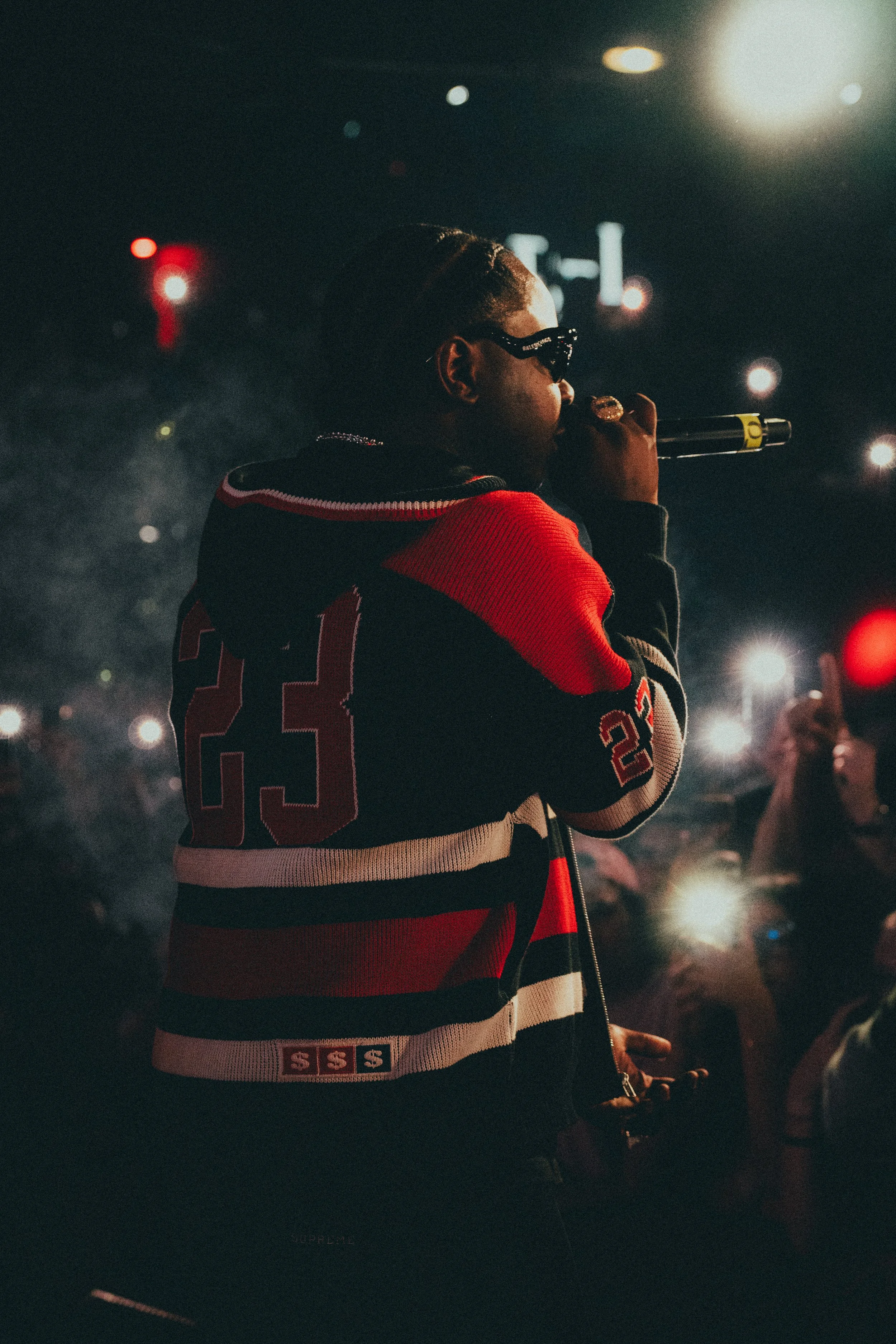 Un artiste chanteur portant un maillot à rayures noirs, rouges et blancs, avec le numéro 23, lors d'une performance en intérieur, entouré de personnes tenant des lumières de téléphone en signe de soutien.
