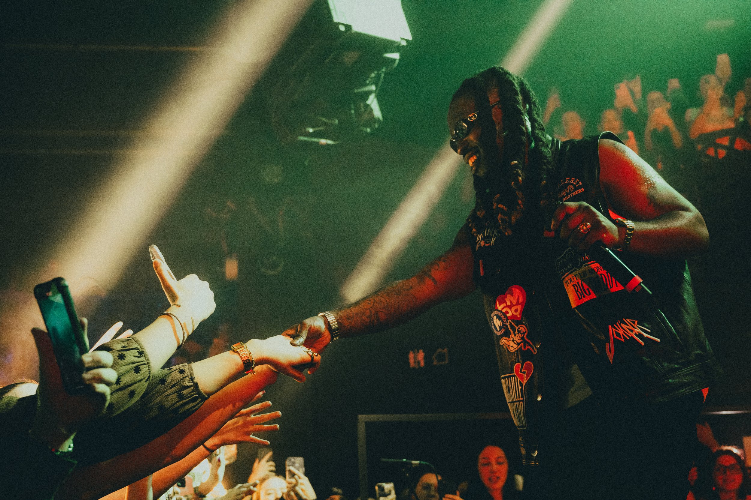Un artiste de musique rap avec des dreadlocks, portant des lunettes noires et un gilet en cuir orné de patches, tend la main à un public enthousiaste lors d'un concert, la scène est éclairée par des faisceaux lumineux en ambiance sombre.