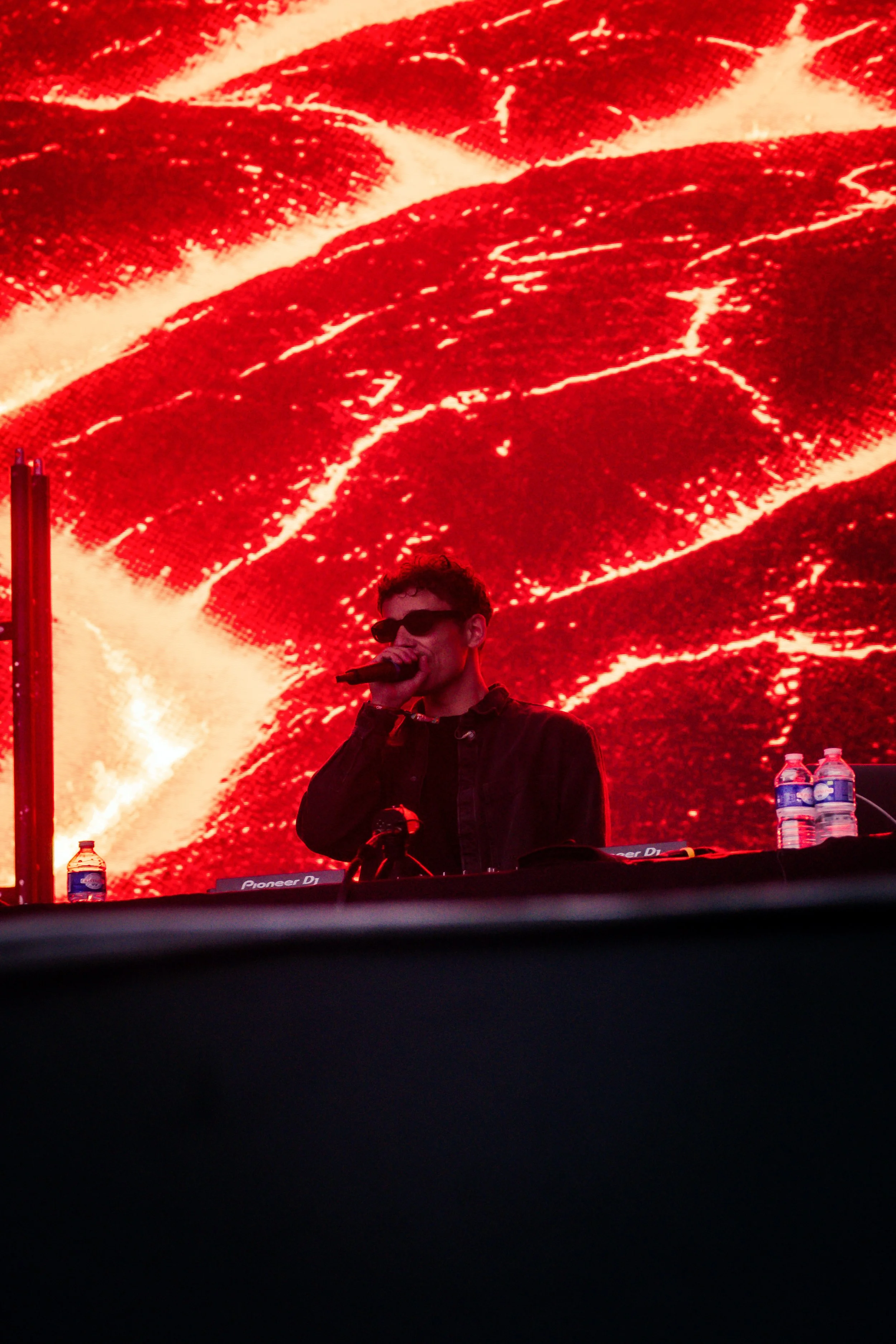 Un DJ portant des lunettes noires joue de la musique sur scène devant un grand écran rouge avec des motifs qui ressemblent à des vagues ou des chaines.