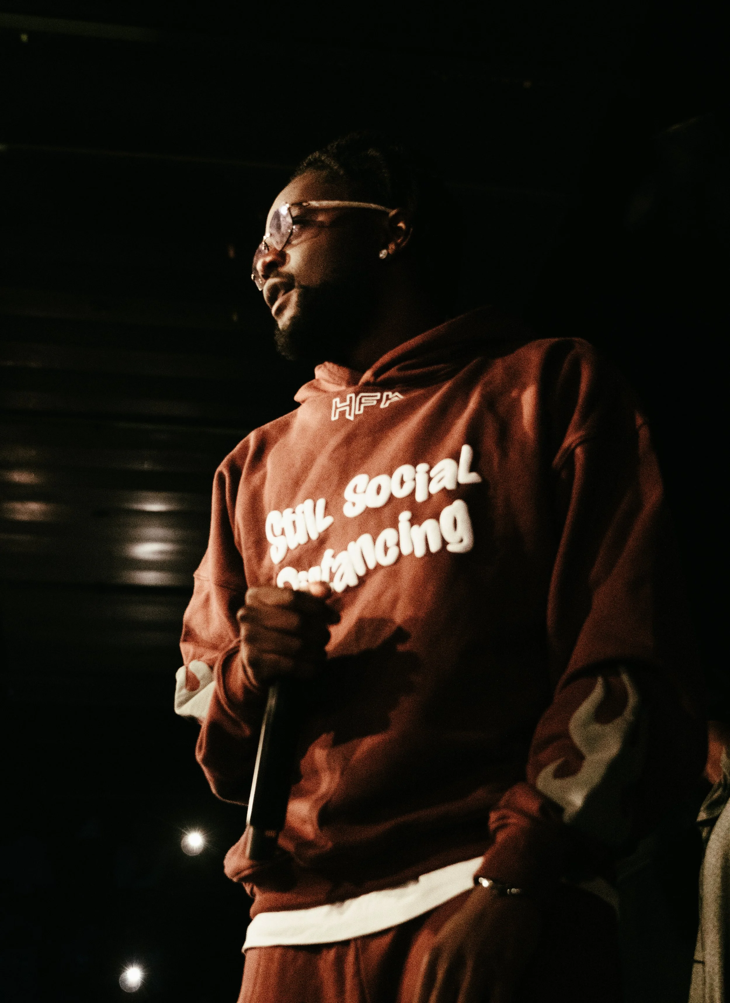 Un homme portant un sweat à capuche marron avec le texte 'Still Social Distancing' écrit dessus, tenant un microphone, dans un environnement sombre.
