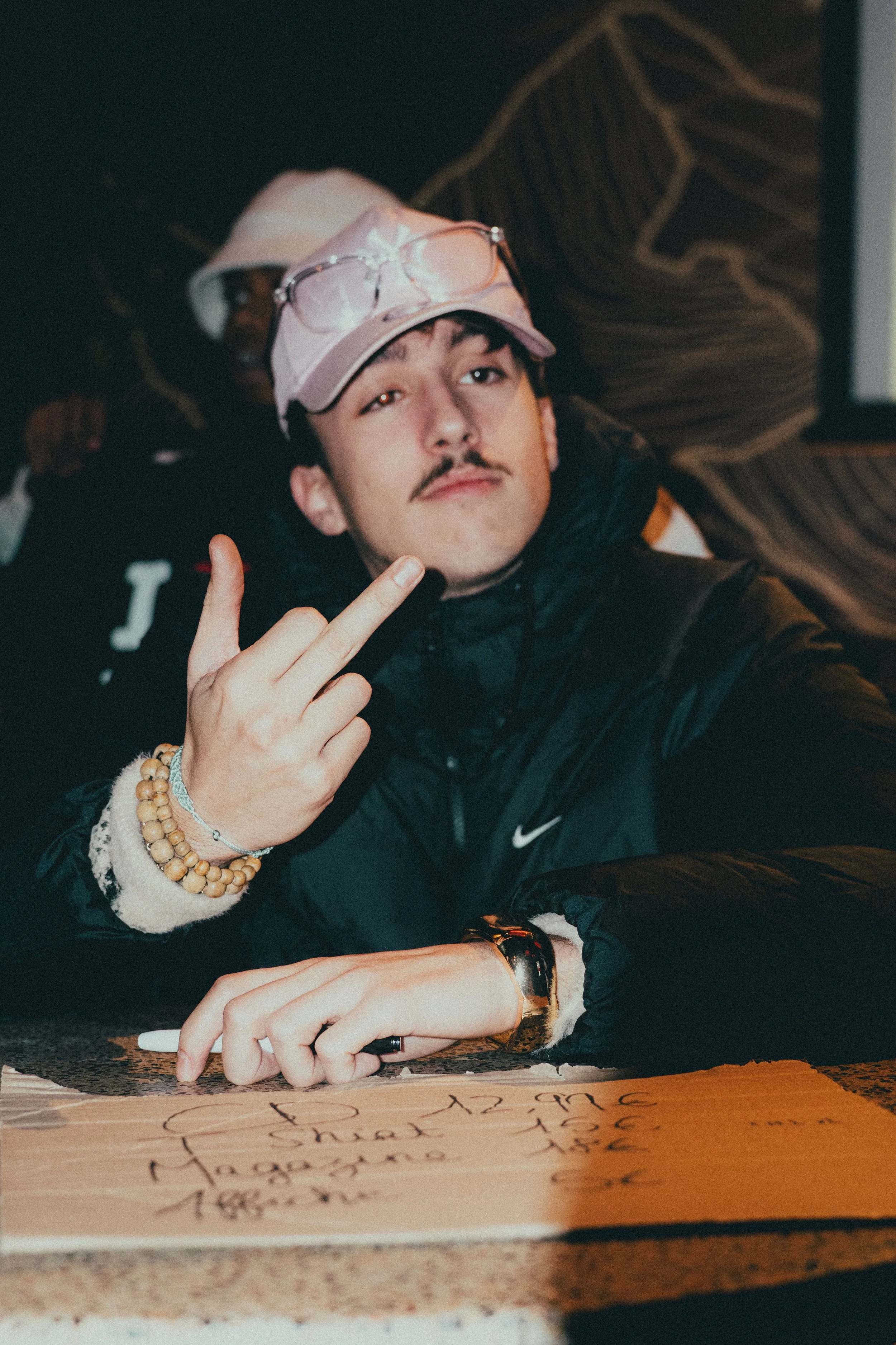 Un jeune homme avec une moustache, portant une casquette rose, un bracelet en bois et une montre dorée, fait un geste avec la main en montrant le majeur. Il est assis à une table en face d'une feuille de papier avec des écrits. Il porte un vêtement n