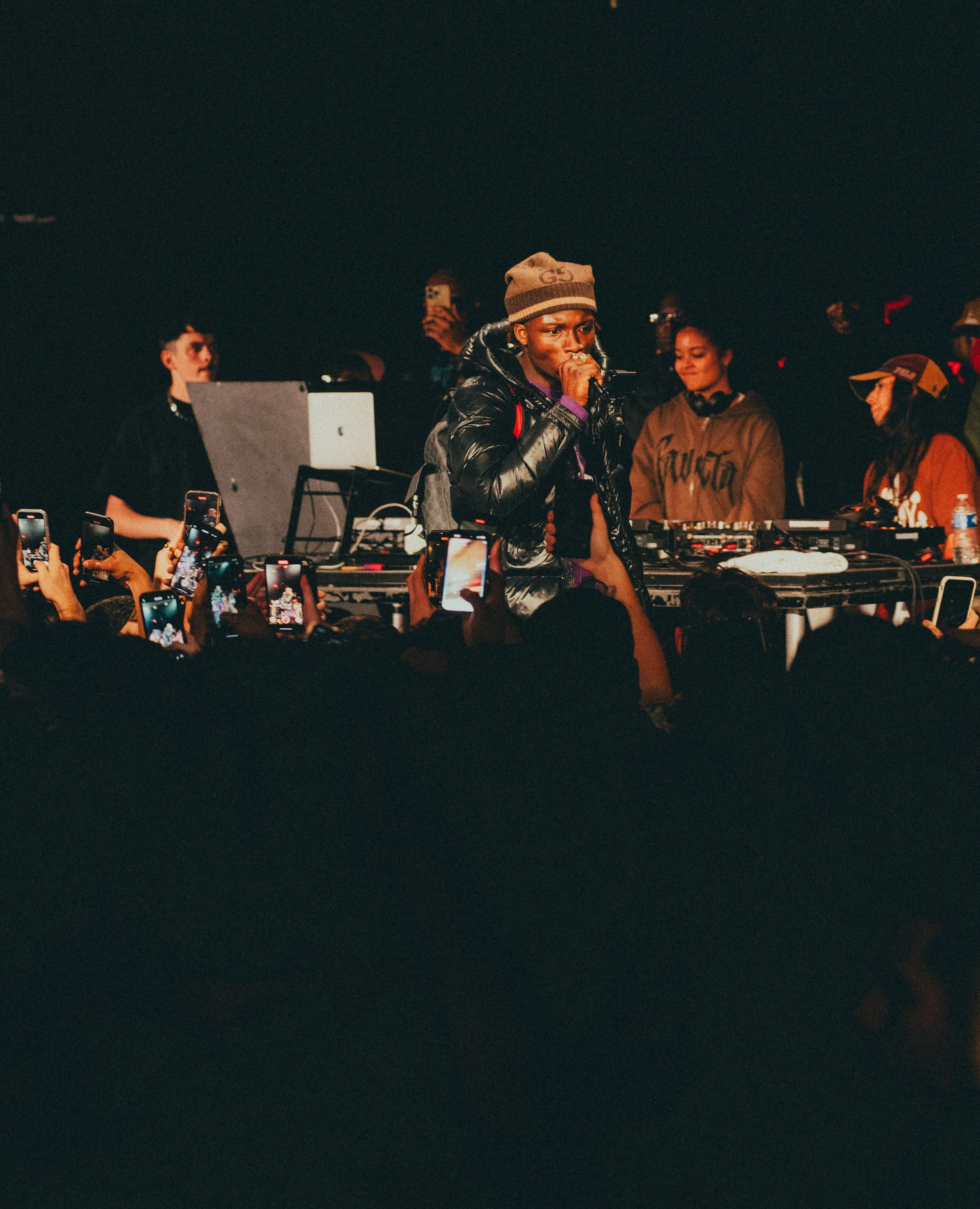 Un DJ sur scène lors d'un concert, entouré de plusieurs personnes, avec un public nombreux prenant des photos et vidéos avec leurs téléphones portables.