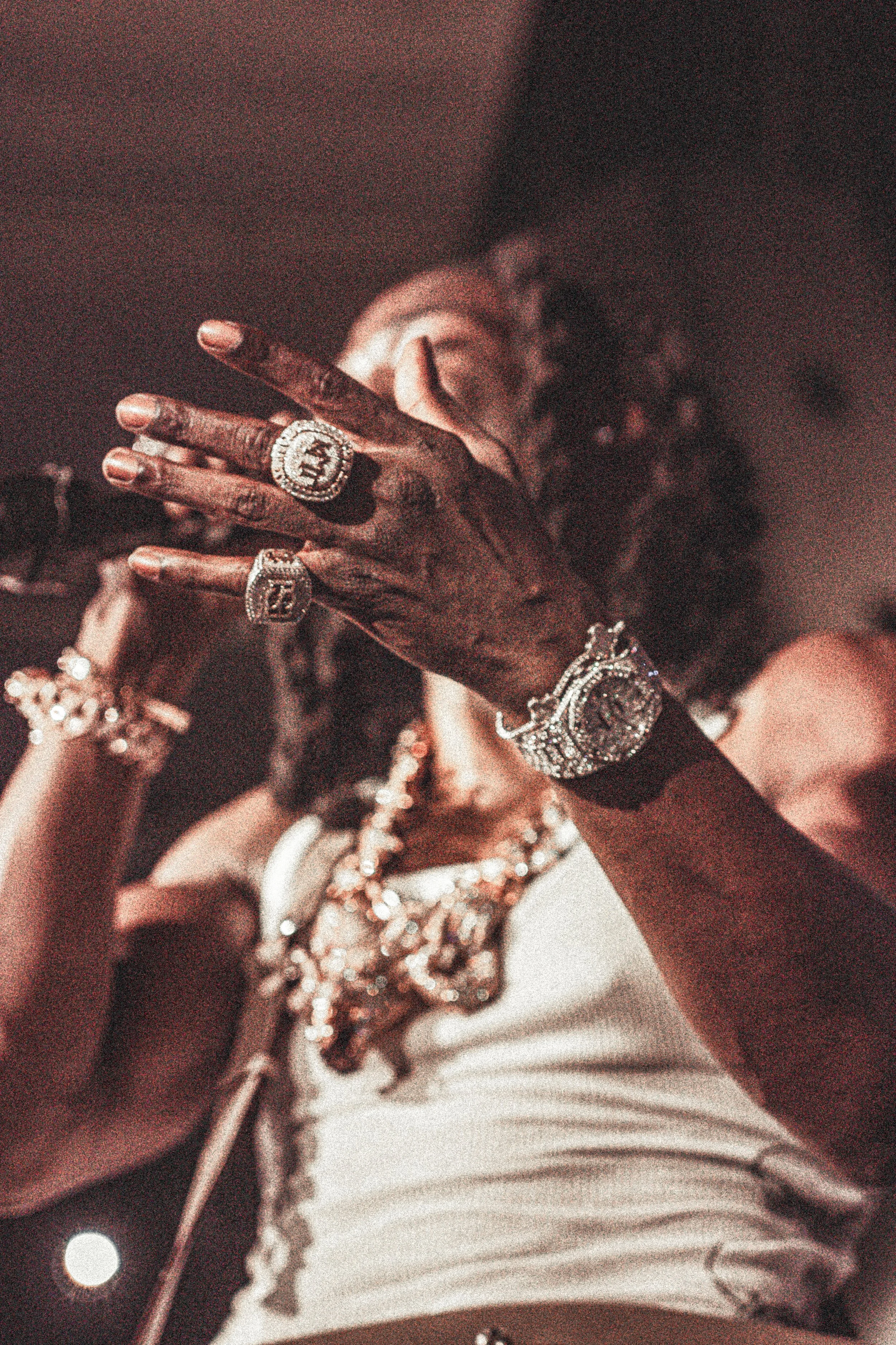 Une personne portant de nombreux bijoux dont des bagues, une montre et un collier, tenant une cigarette, avec des cheveux bouclés.