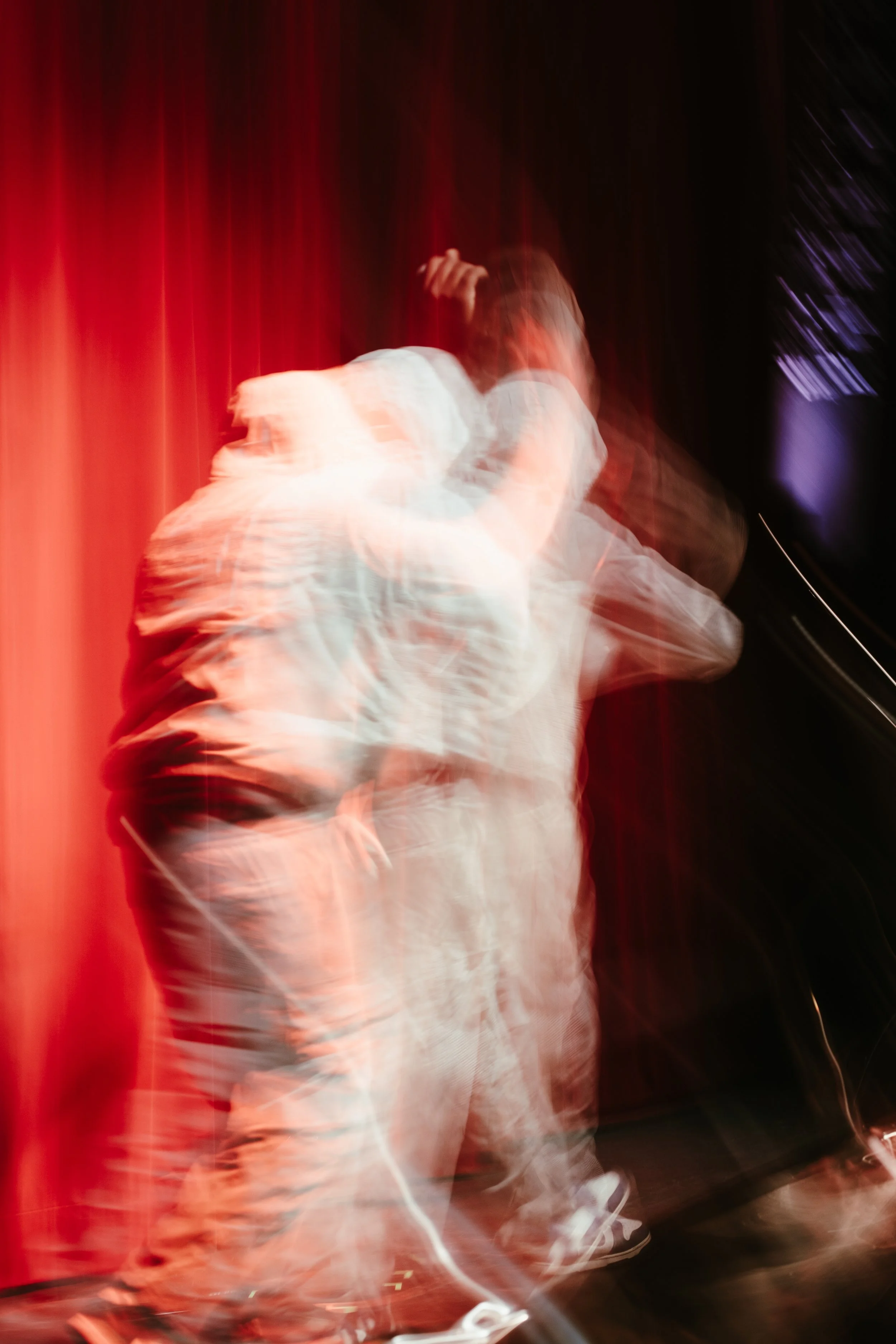 Photo floue d'une personne qui danse sur scène, avec un rideau rouge en arrière-plan, créant un effet de mouvement.