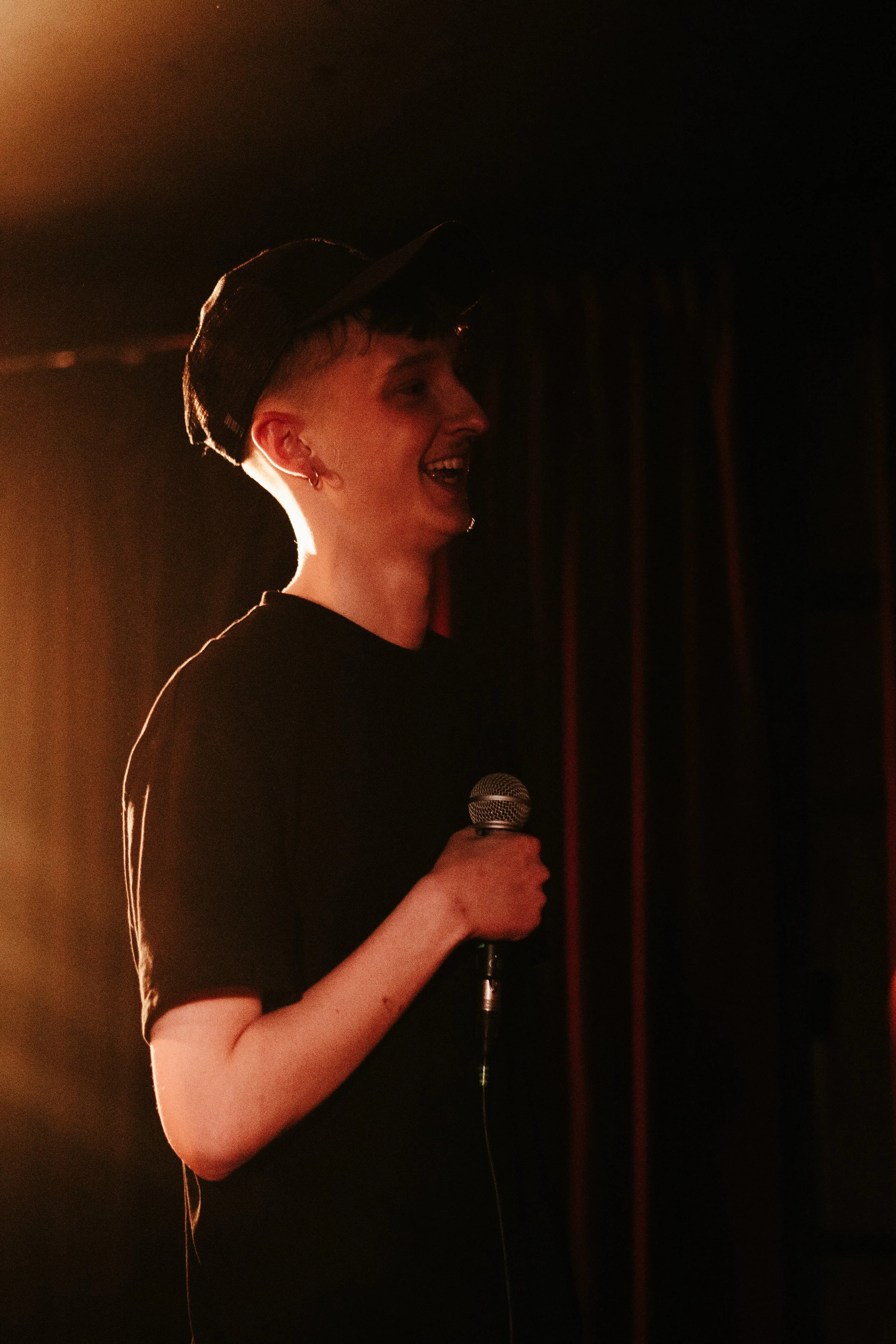 Une jeune femme souriante tient un micro lors d'une performance ou d'une présentation dans une scène sombre avec un éclairage chaud.