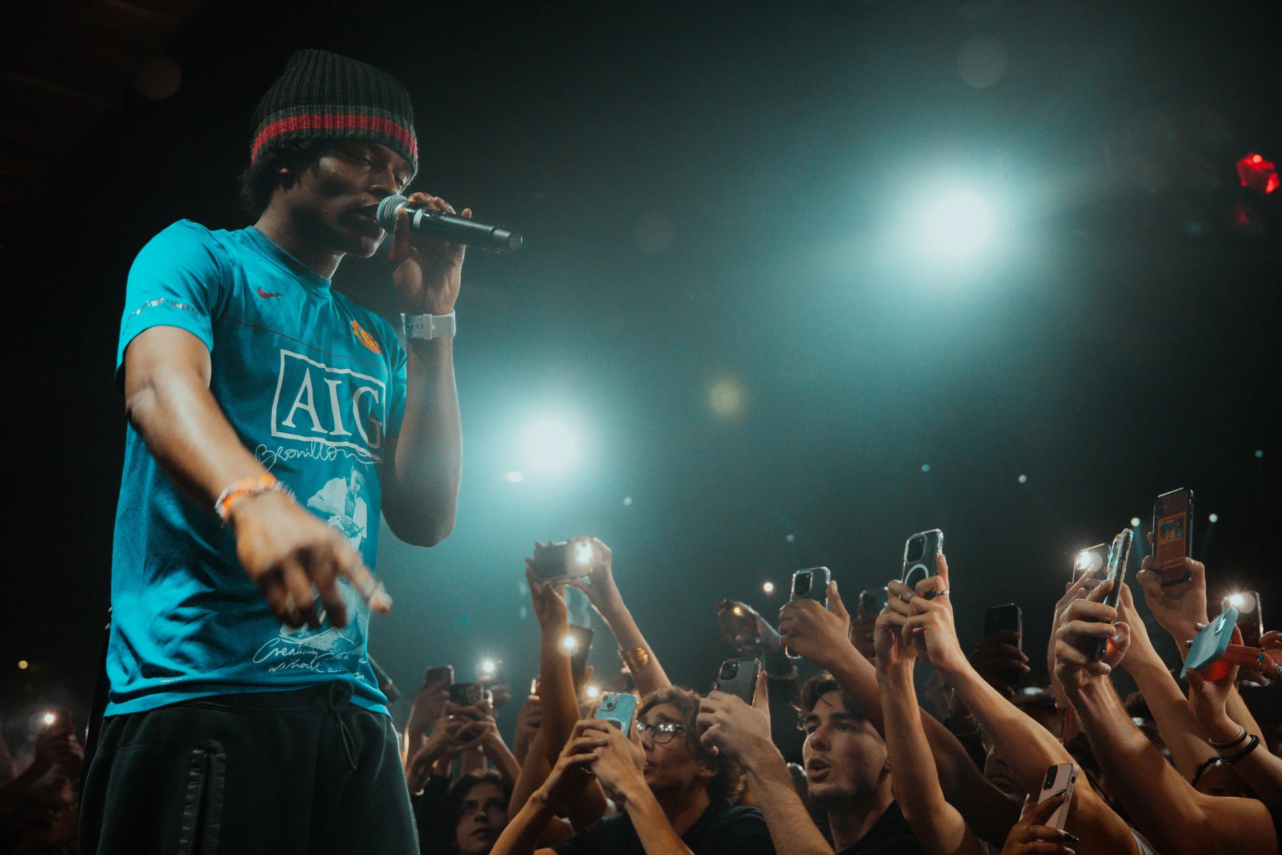 Un homme artiste chante en direct lors d'un concert devant une foule captivée, avec beaucoup de personnes utilisant leurs téléphones portables pour prendre des photos ou vidéos.