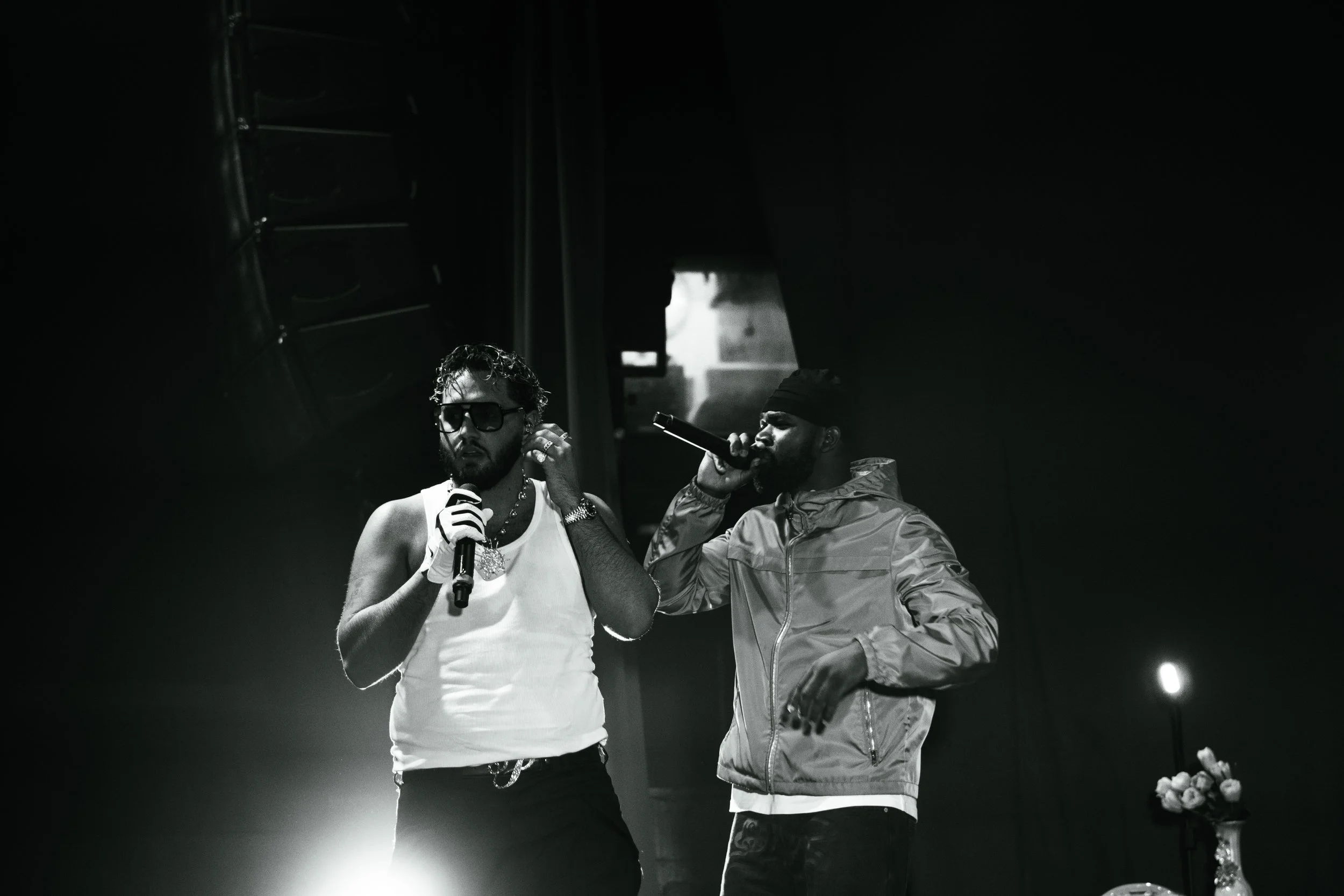 Deux hommes chantent avec des microphones sur scène, l'un porte des lunettes de soleil et un tank top blanc, l'autre porte une casquette et une veste