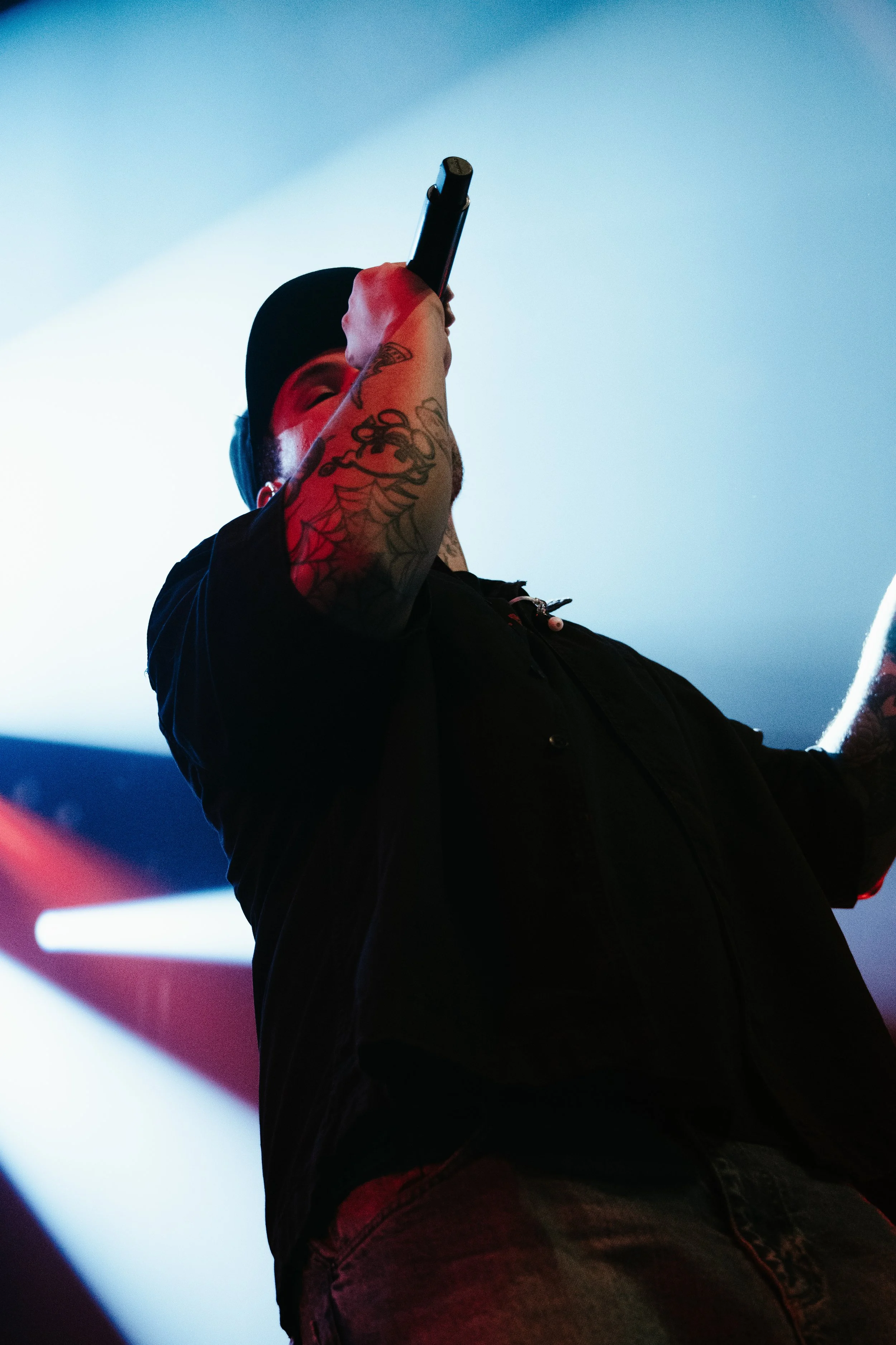 Un homme tatoué portant une casquette noire chante ou parle dans un microphone sur scène, avec un fond de lumière bleue et rouge.