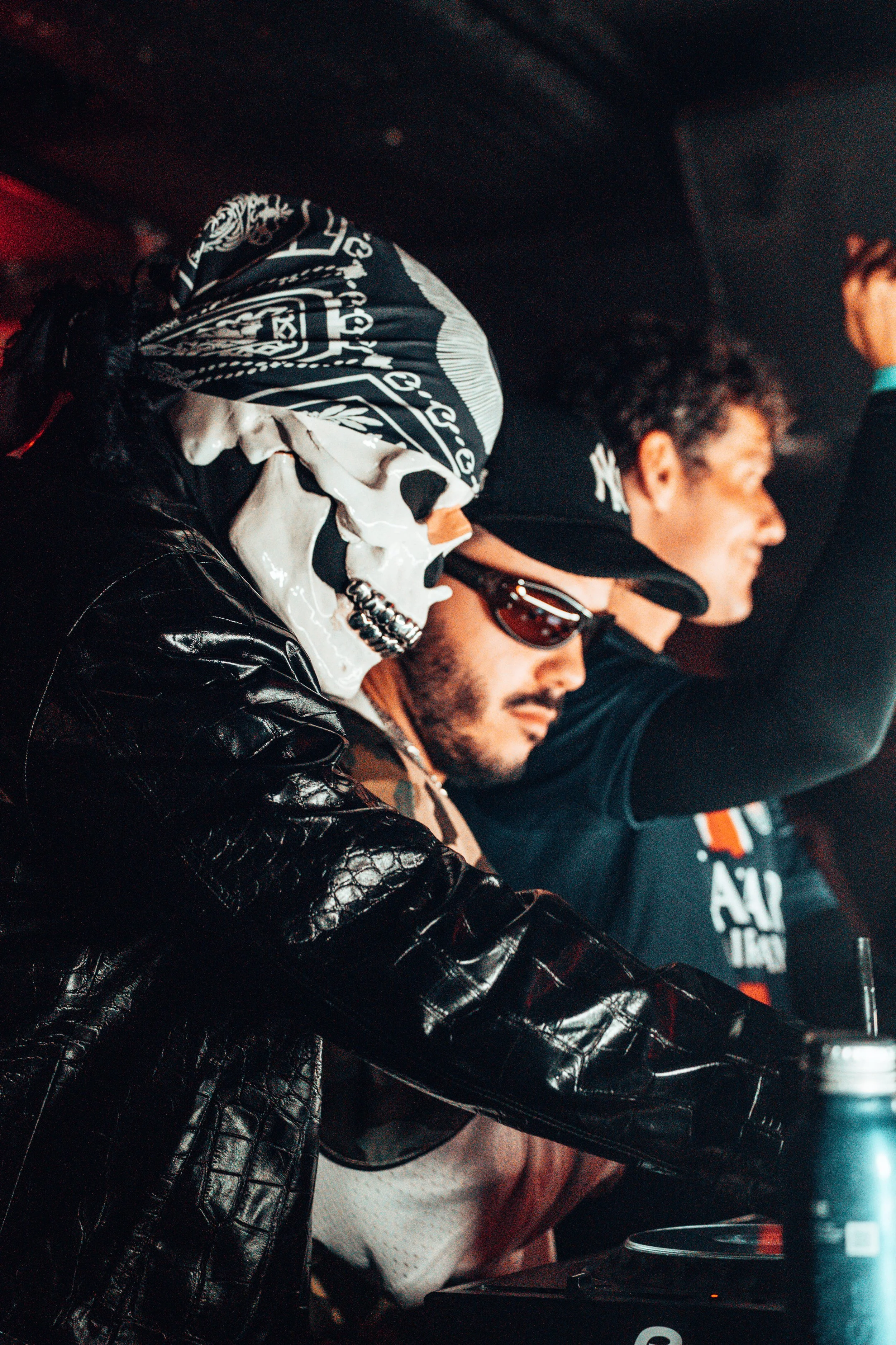 Trois hommes dans une scène sombre, l'un portant un masque en forme de crâne et un bandana, un autre portant des lunettes de soleil et une casquette, le troisième portant un t-shirt noir.