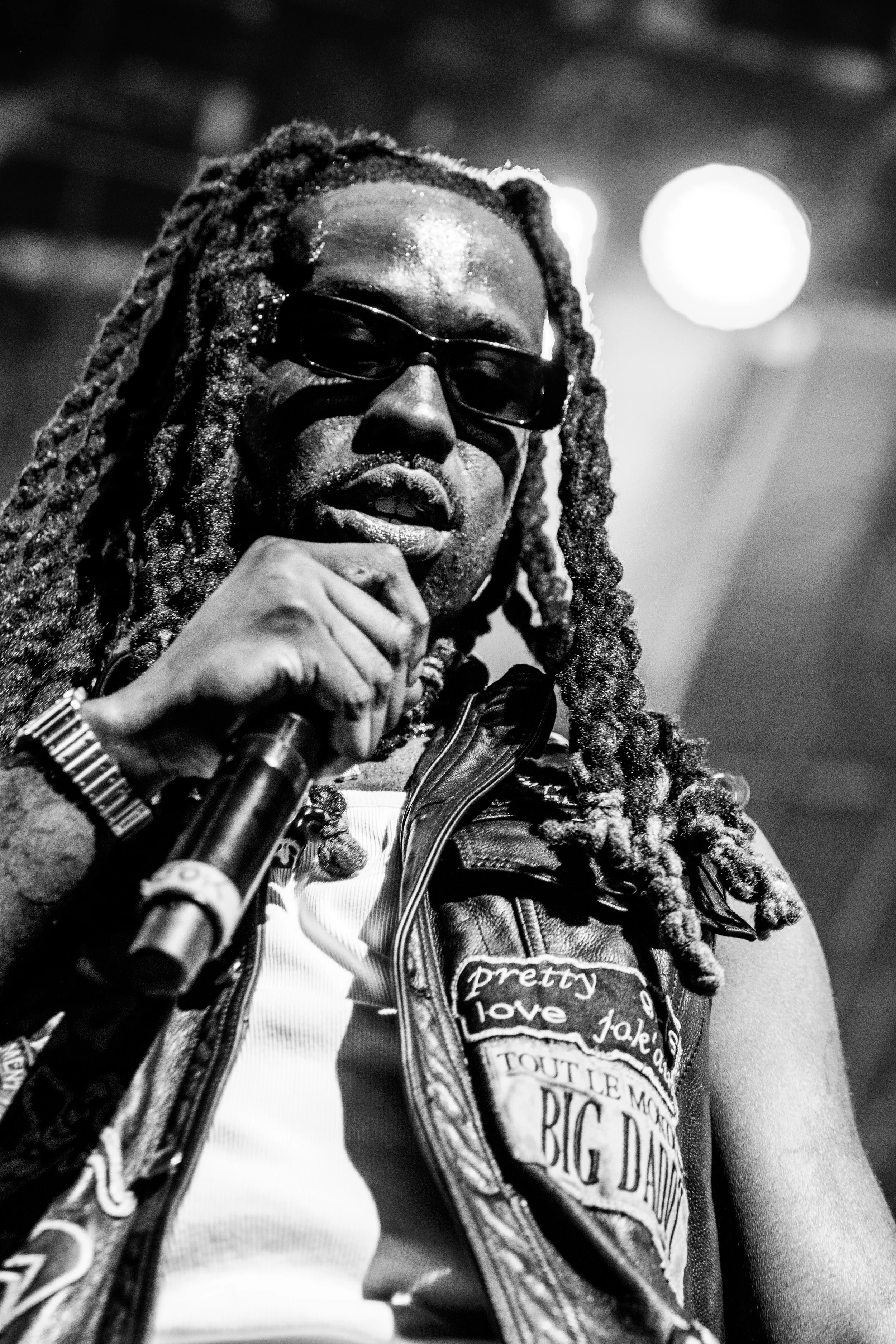 Un homme avec de longs dreadlocks, porteur de lunettes de soleil, tenant un microphone, portant un gilet en cuir avec différents patches, dans un environnement éclairé.