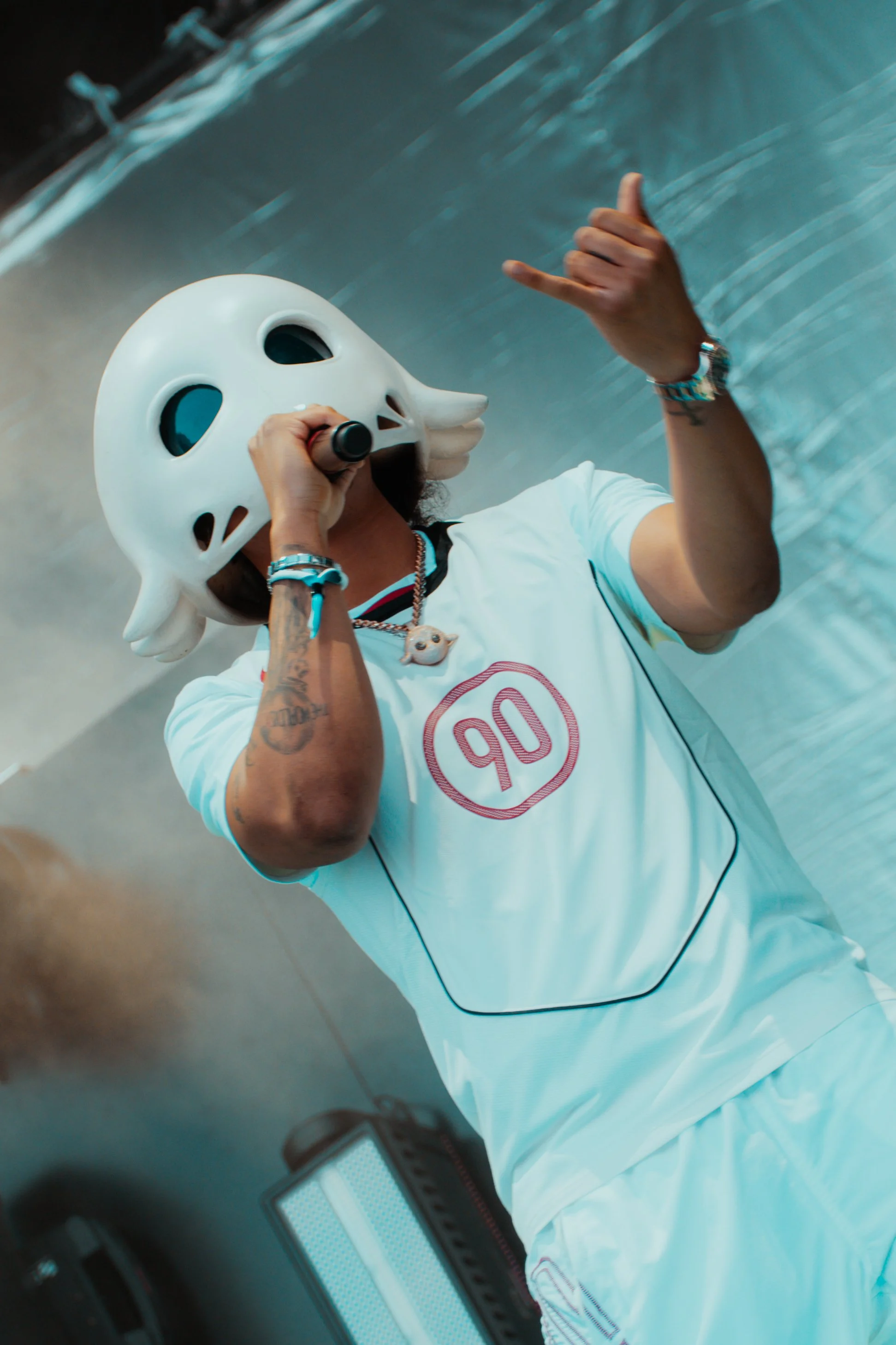 Personne portant un casque de masque de fantôme blanc, chantant ou parlant dans un micro, tenant la main en signe de shaka, portant un t-shirt blanc avec un logo rouge, à côté d'une caisse de musique.