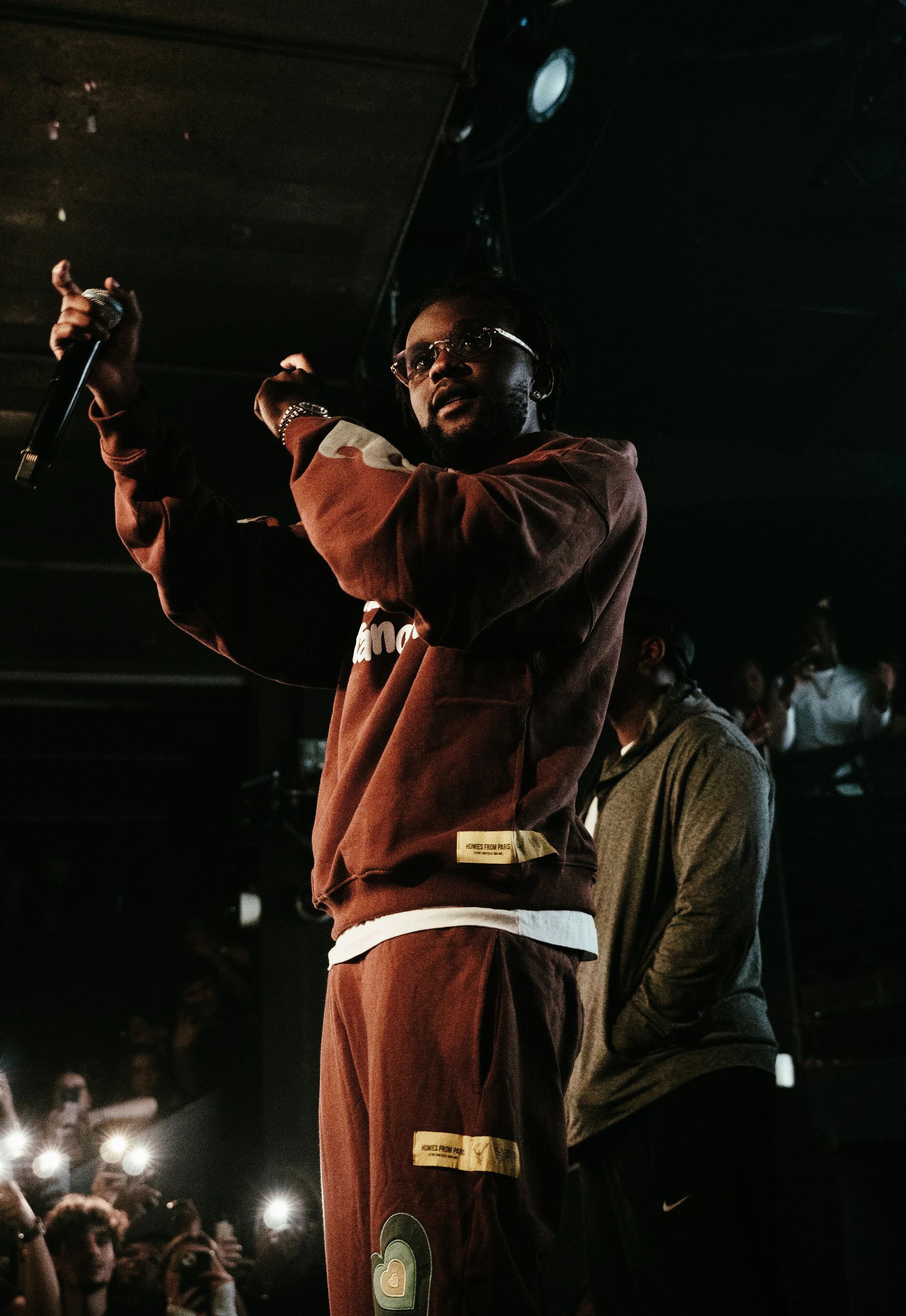 Un homme tenant un micro sur scène lors d'une performance, avec un public qui l'éclaire avec leurs téléphones. L'ambiance est sombre, avec un éclairage concentré sur lui.